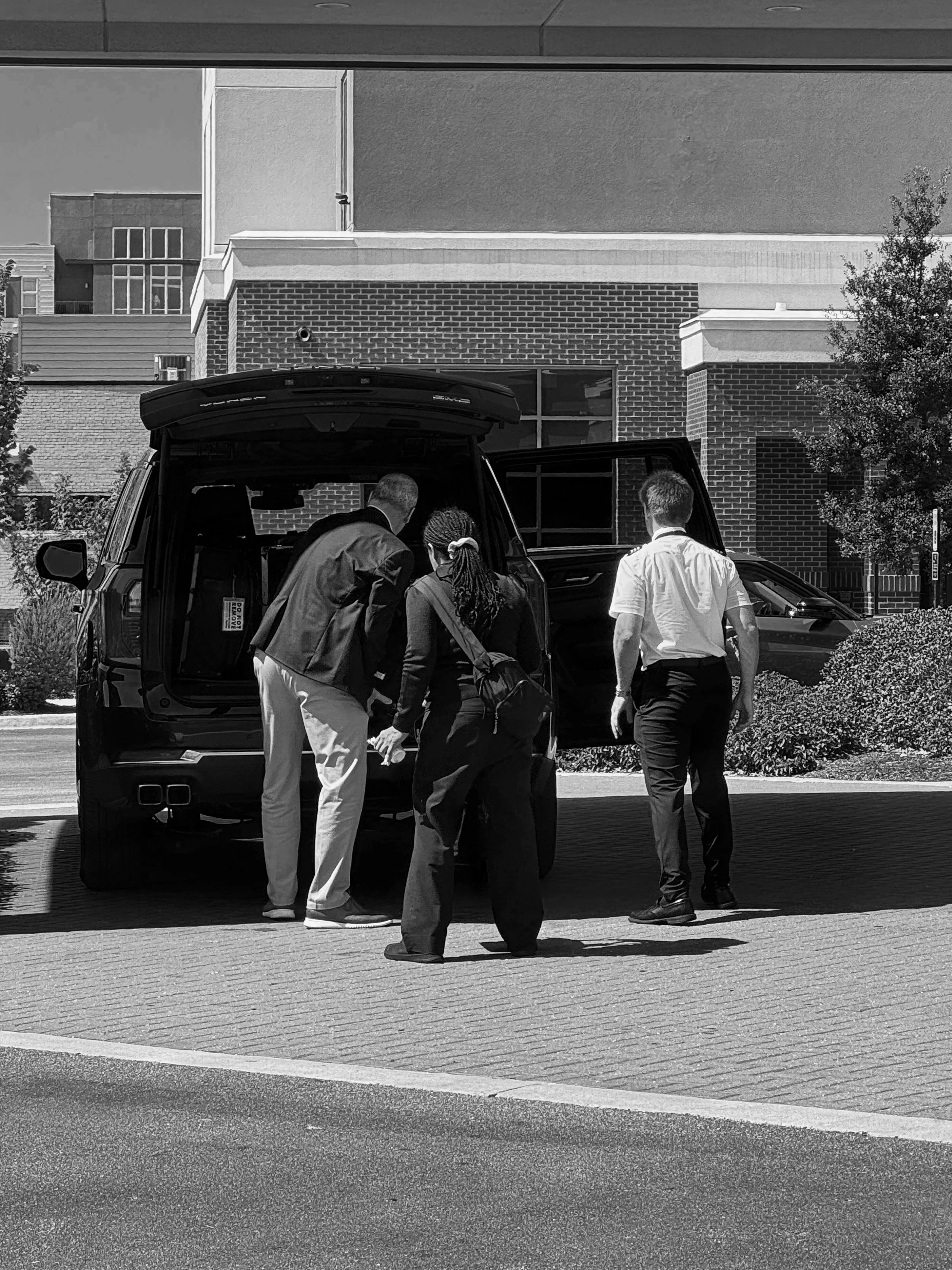 Three people, dressed in business attire, are gathered around the open trunk of a black SUV, parked outside a modern building with brick and stucco exterior, in an urban area. One person is leaning into the trunk, and the other two are standing nearby.