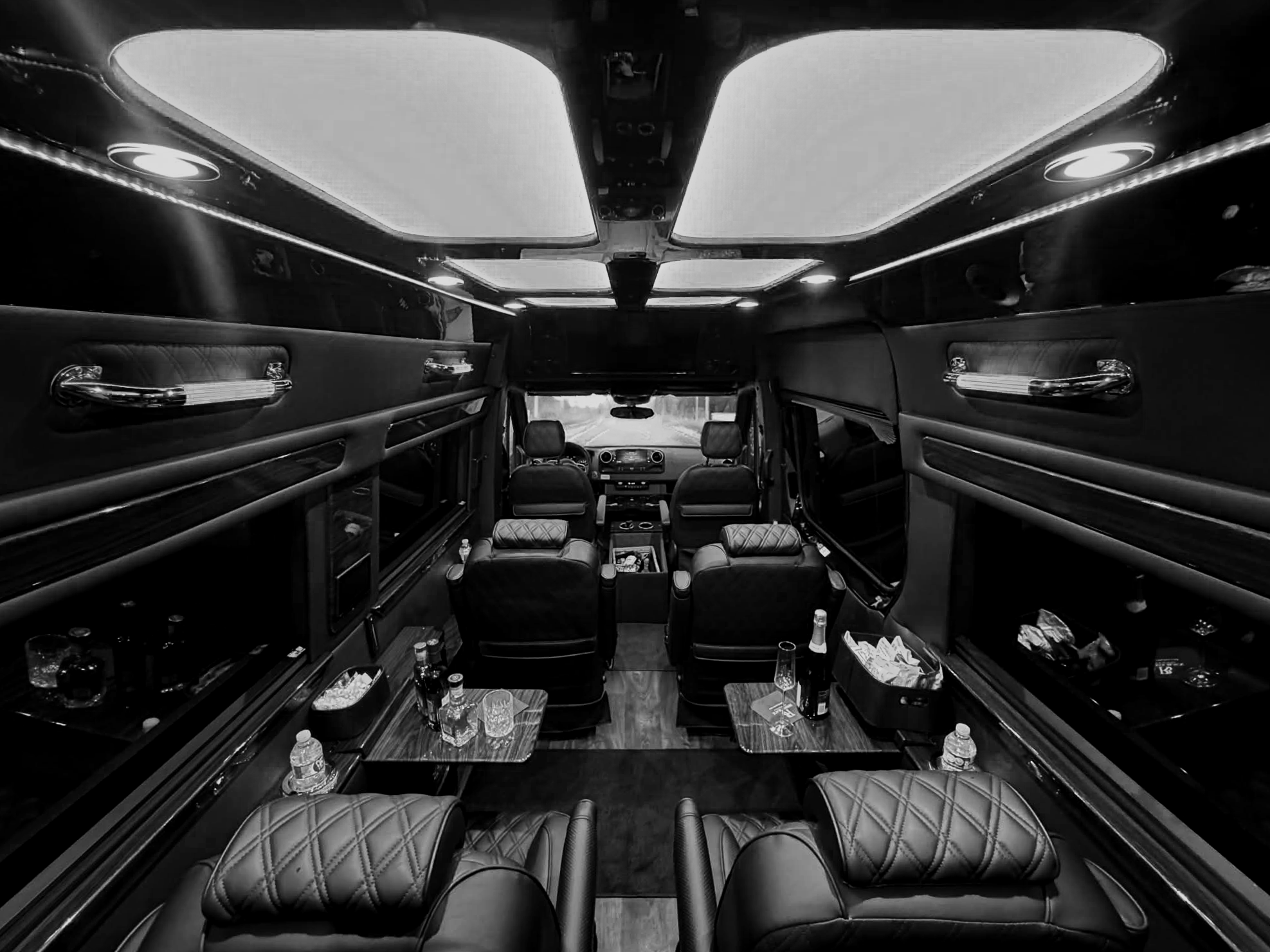 The interior of a luxury limousine with leather seats, tables with drinks, and a row of overhead compartments.