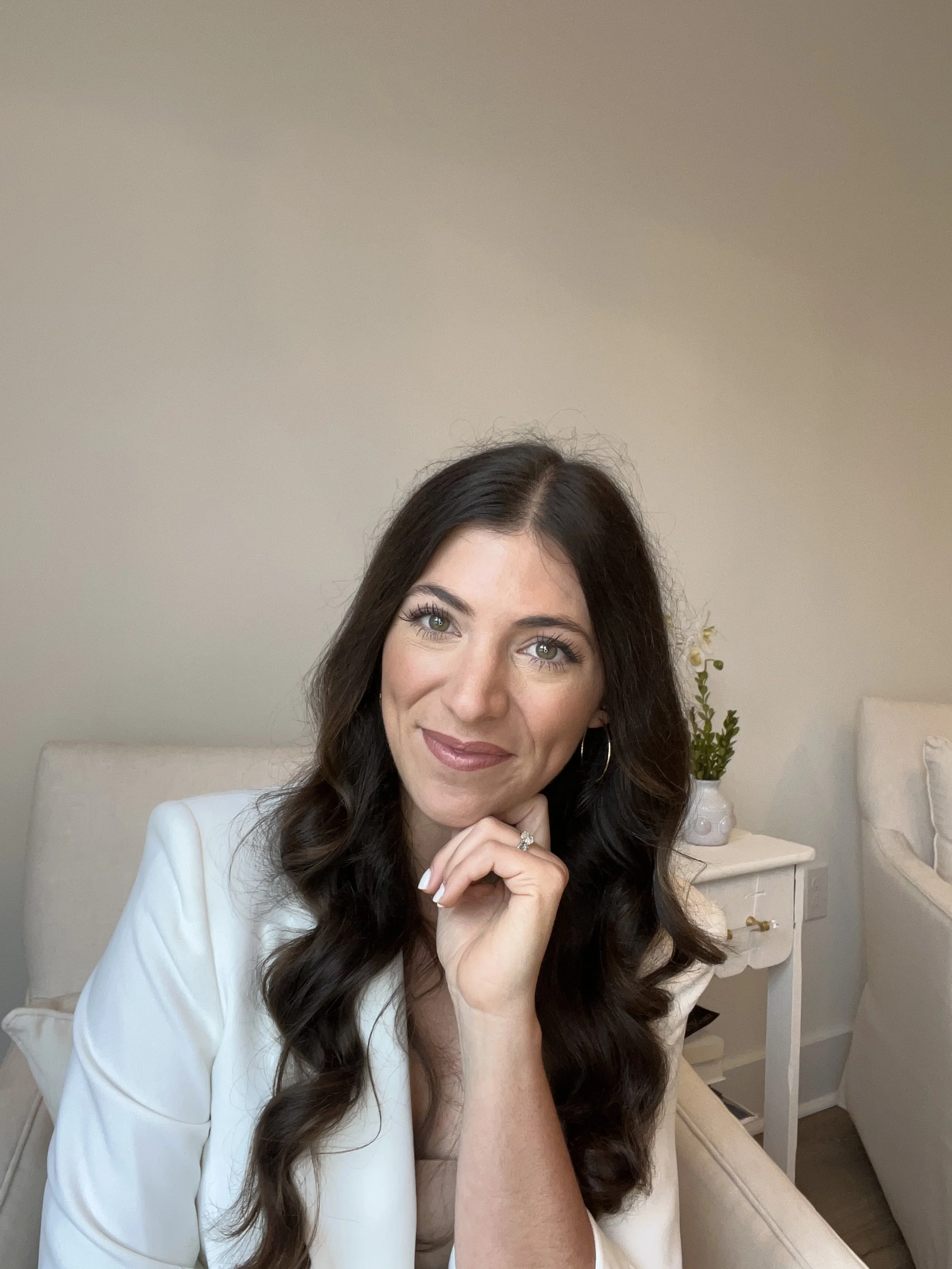 A woman with long, dark, wavy hair and blue eyes, wearing a white blazer, sitting on a beige sofa with a white and green plant in the background.