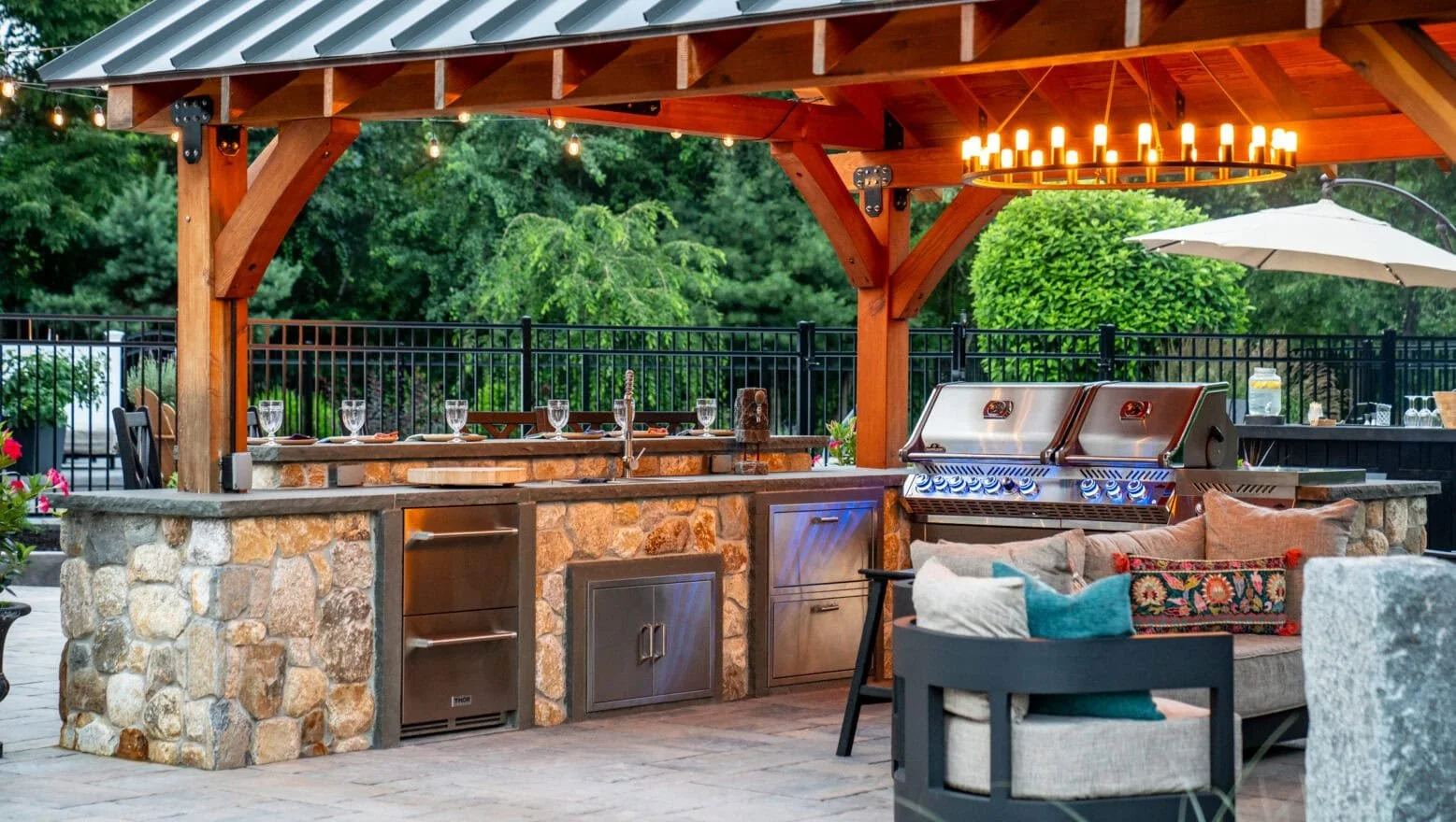 Outdoor kitchen and seating area with a stone counter, built-in grill, and wood pergola, lit by a chandelier and string lights, surrounded by trees and a black fence.