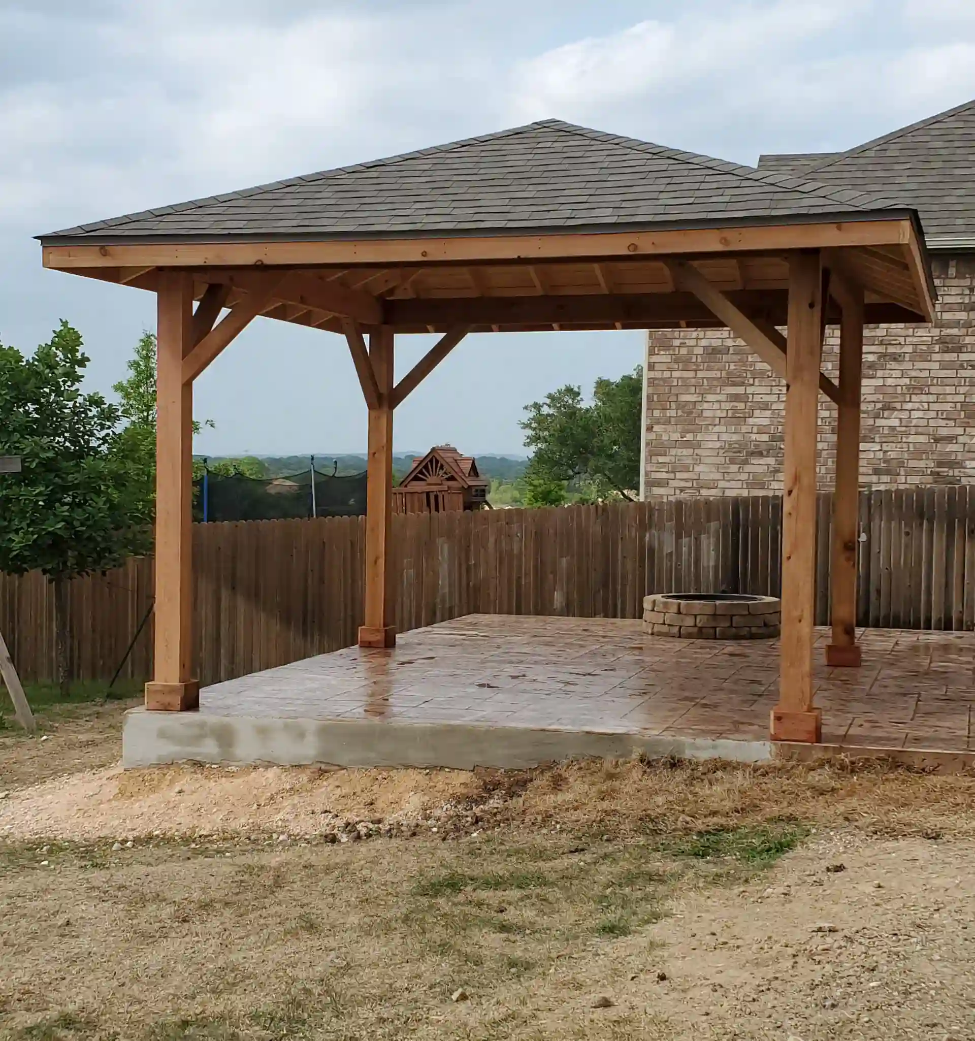 A backyard patio with a wooden gazebo, stamped concrete floor, brick fire pit, surrounded by a wooden fence, with trees and a house in the background.