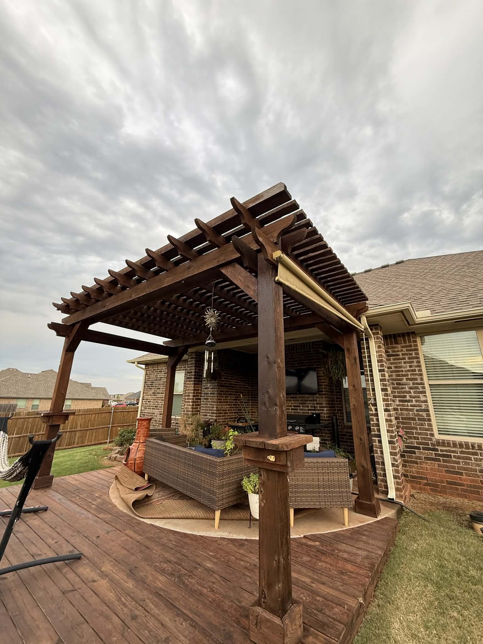 Backyard patio with a wooden pergola, wicker sofa, potted plants, and outdoor decor against a brick house with windows.