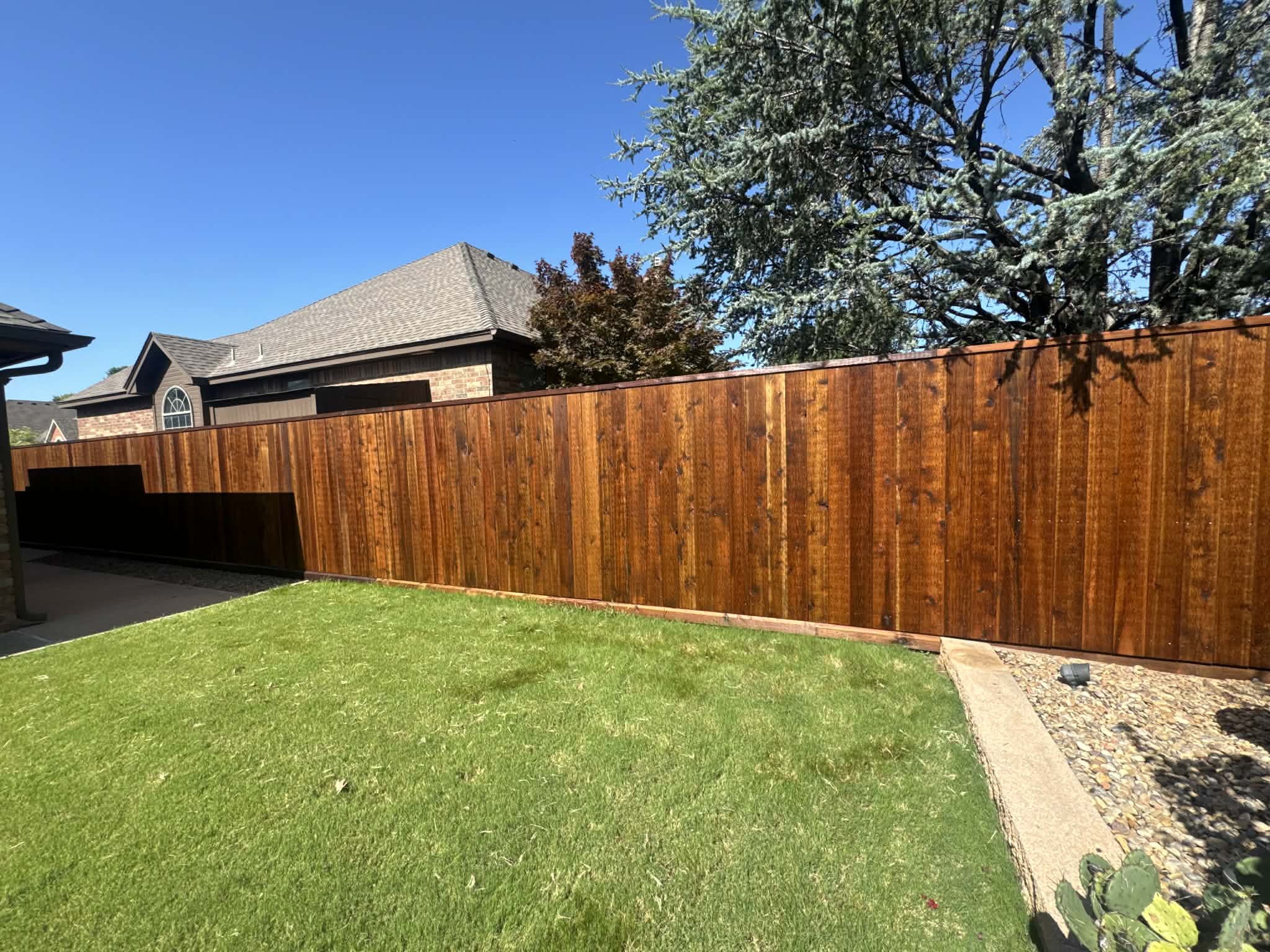 View of a backyard with a green lawn, a wooden fence, a small gravel area, and part of a house with a brick wall and a window, under a clear blue sky.