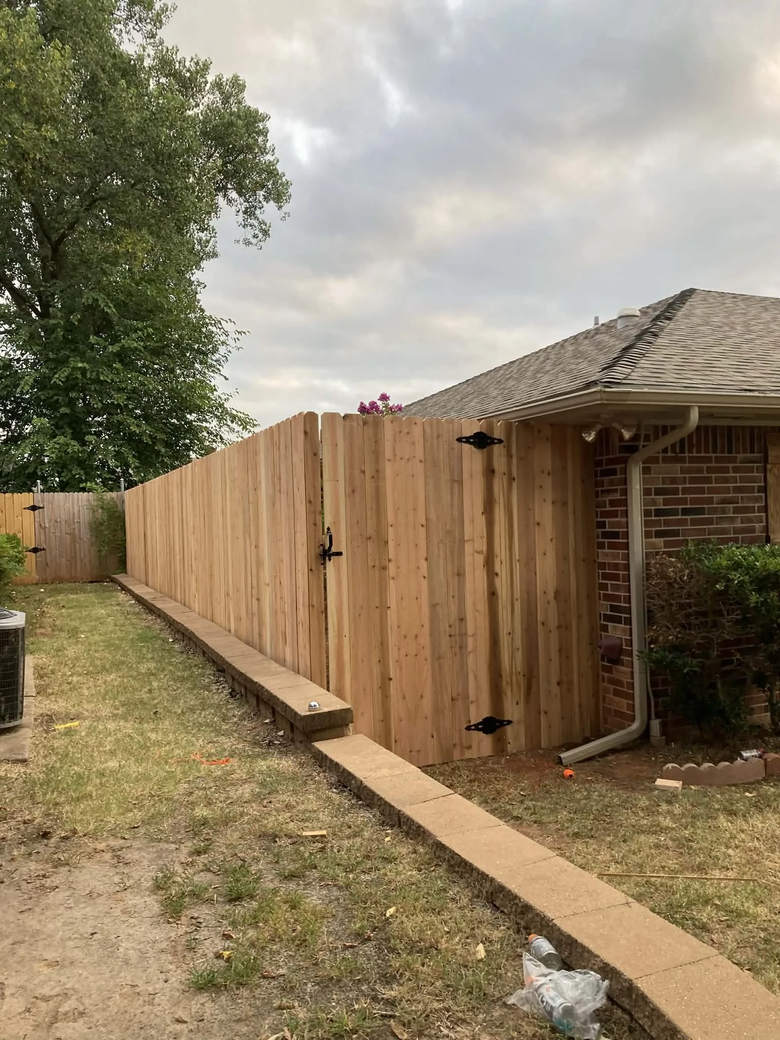 New wooden privacy fence installed adjacent to a brick house with some gardening bushes, on a grassy yard under a cloudy sky.