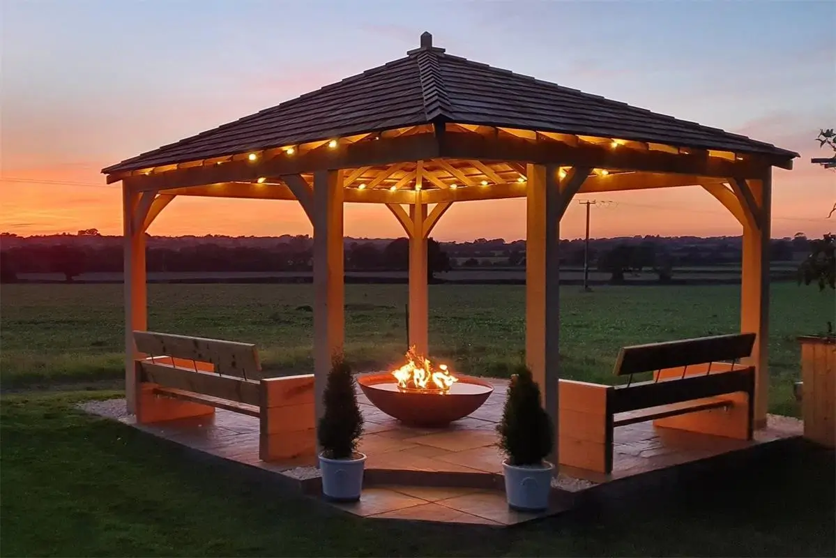 A wooden gazebo with string lights and a fire feature at sunset, surrounded by potted plants and an open grassy field.