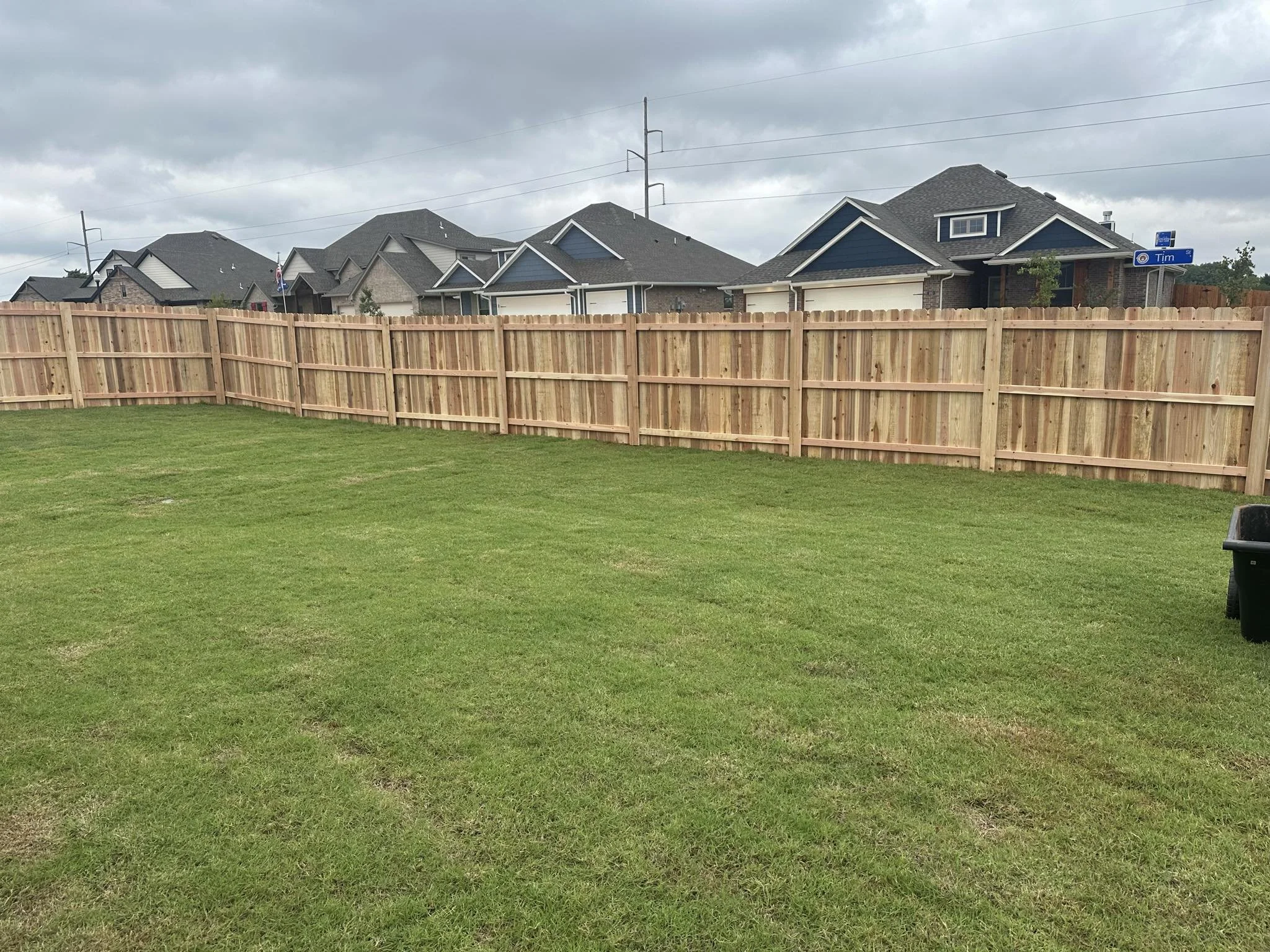 A backyard with a freshly built wooden fence, green grass, and a cloudy sky. Several houses are visible in the background.