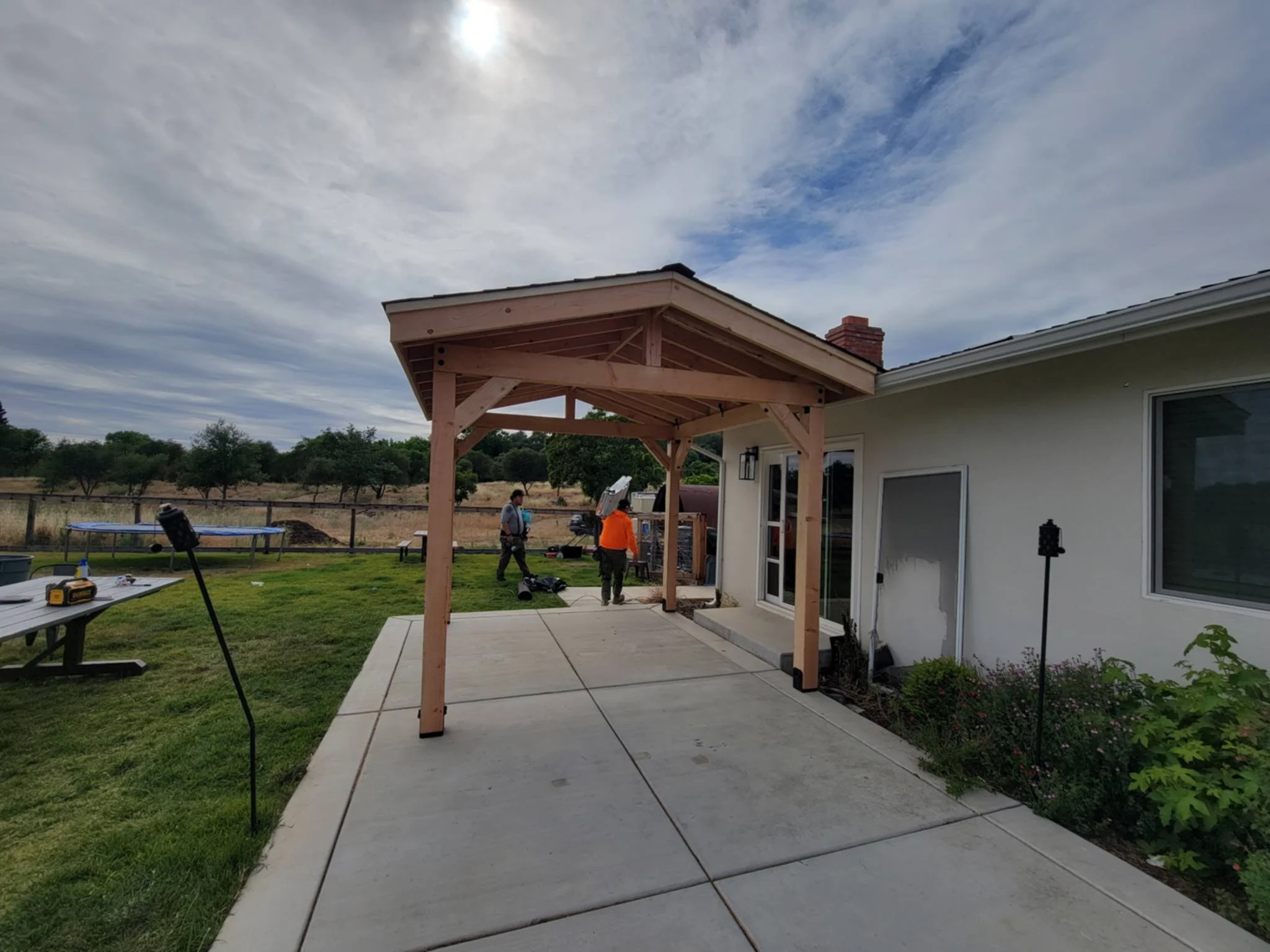 Construction of a wooden porch canopy attached to a house on a concrete patio, with workers in the background and tools on a nearby table.