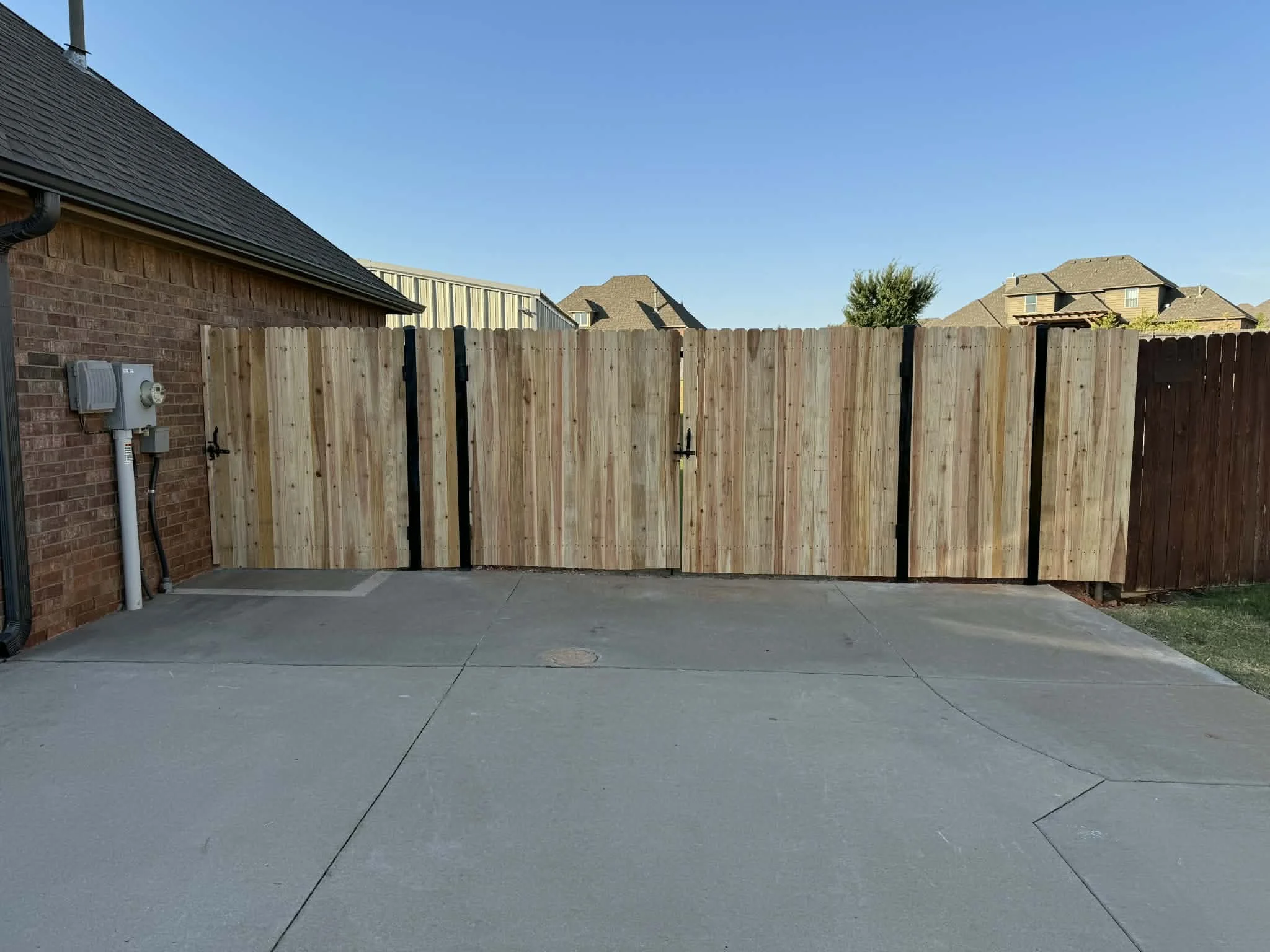 Backyard with a wooden fence gate, brick house wall, concrete patio, and neighboring houses in the background under a clear blue sky.