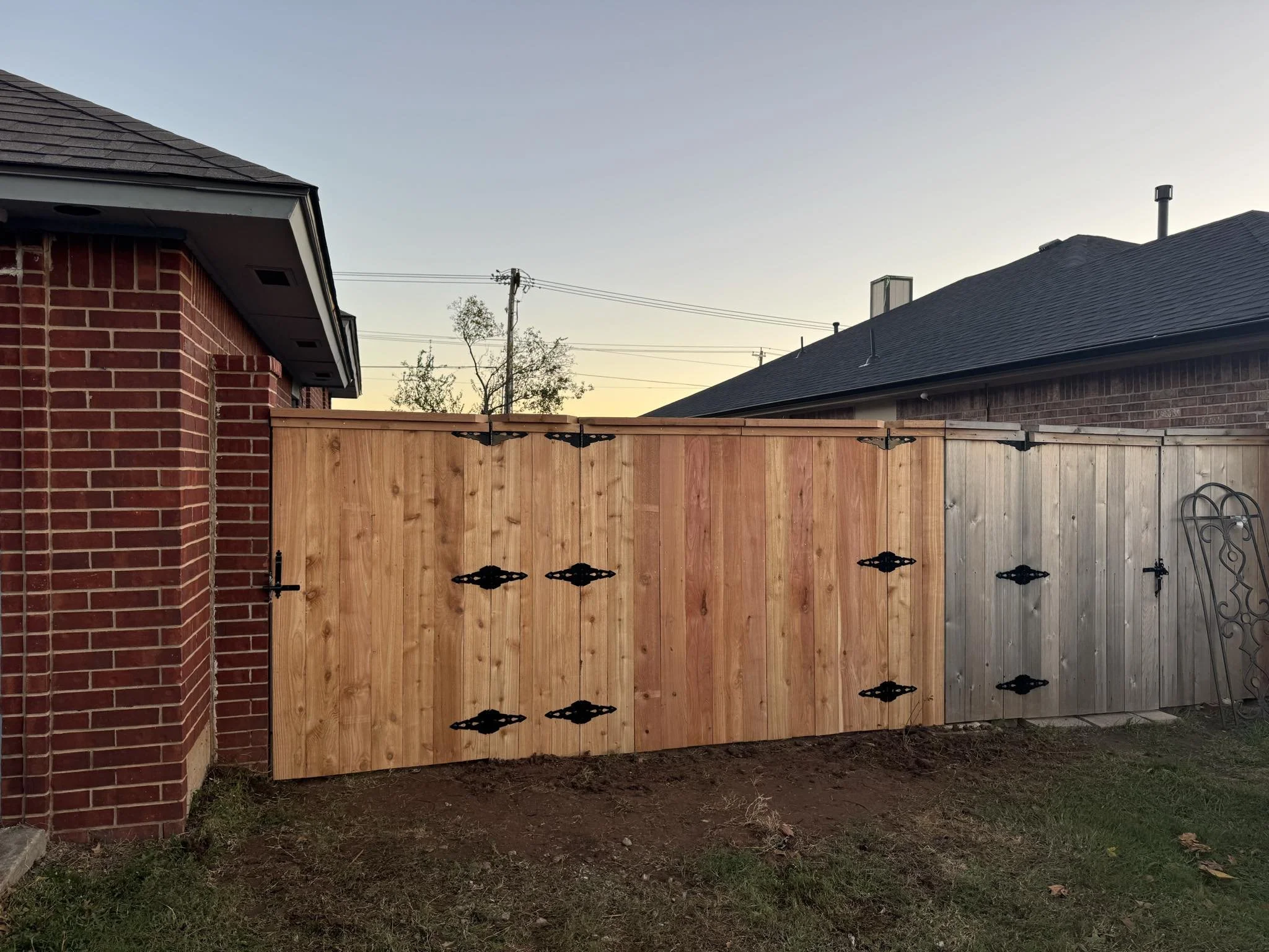 A new wooden fence with black decorative hinges installed between two houses, one with red brick exterior and the other with darker shingles, under a clear sky during sunset.
