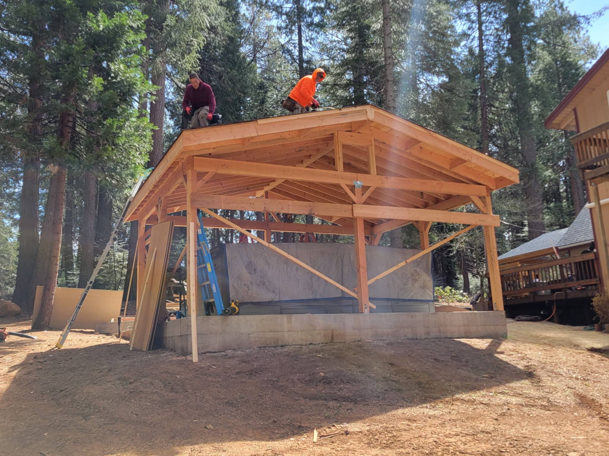 Two construction workers on the rooftop of a wooden building under construction, surrounded by trees and other structures.