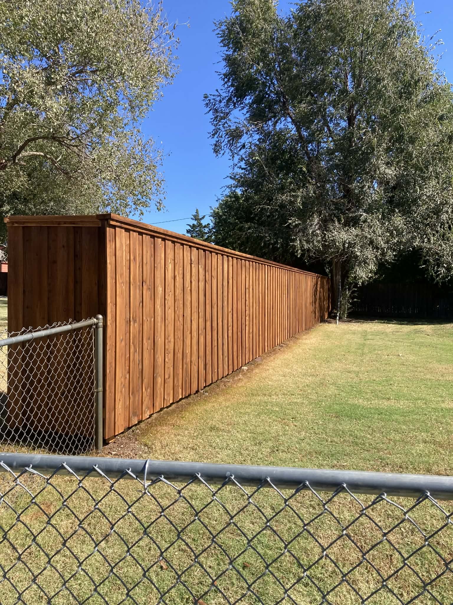A new wooden privacy fence runs along a grassy backyard, with a chain-link fence in the foreground and large trees in the background under a clear blue sky.