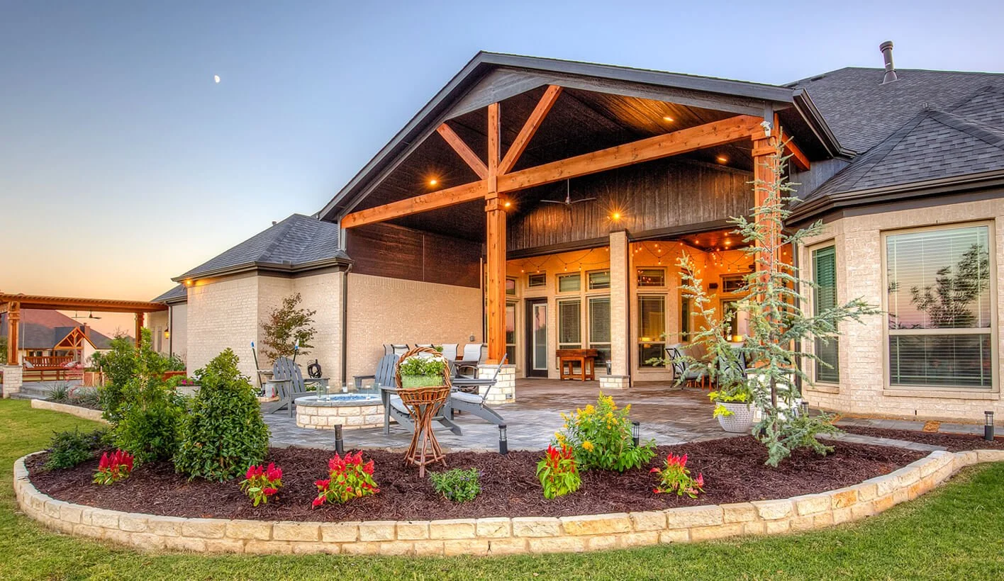 Backyard patio with outdoor seating, fire pit, landscaped flower bed, and covered porch of a house during sunset.