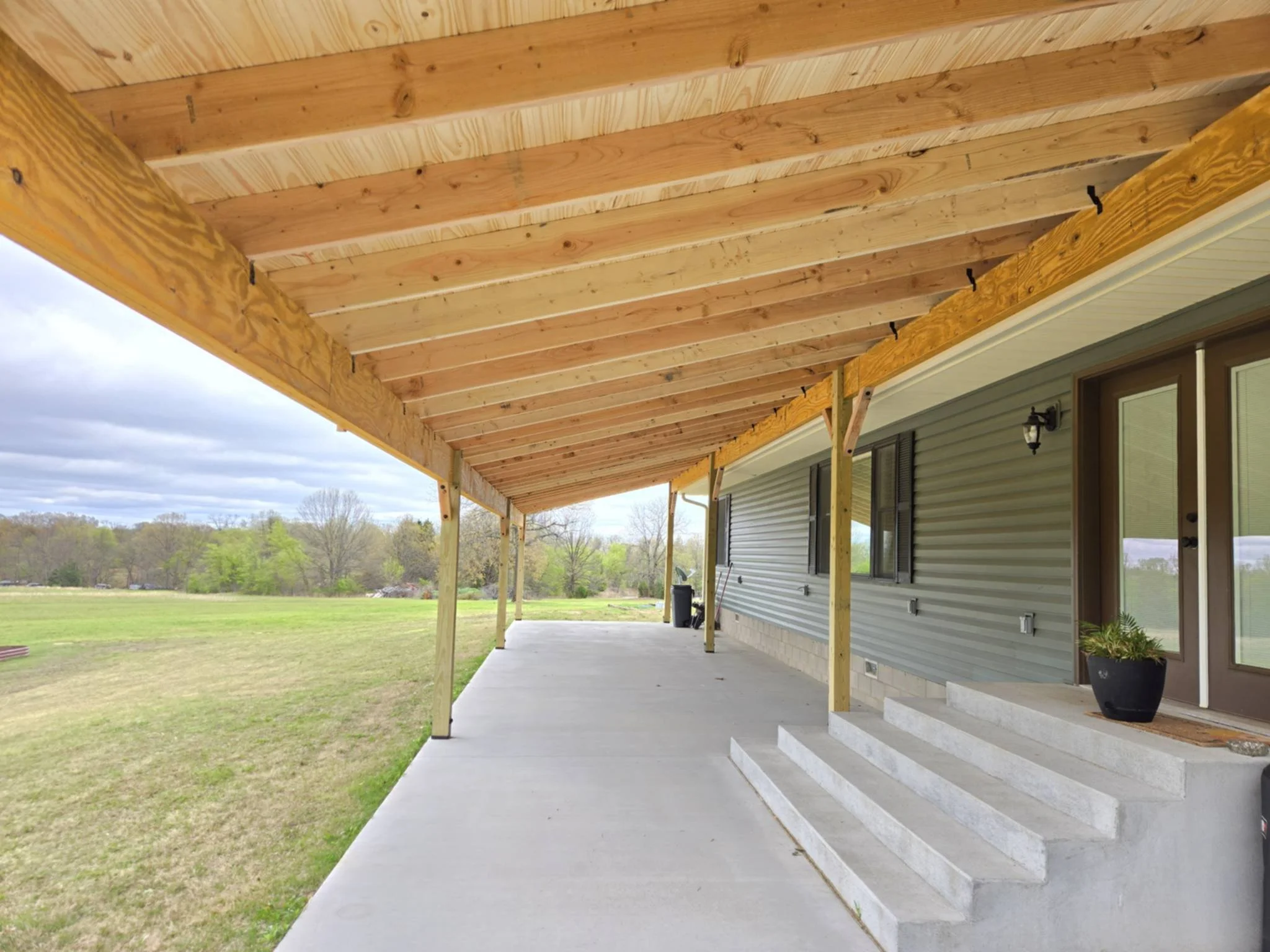 View of a covered porch with a concrete floor, steps, wooden beams, and a metal-clad house exterior with windows and a door, overlooking a grassy field with trees in the background.
