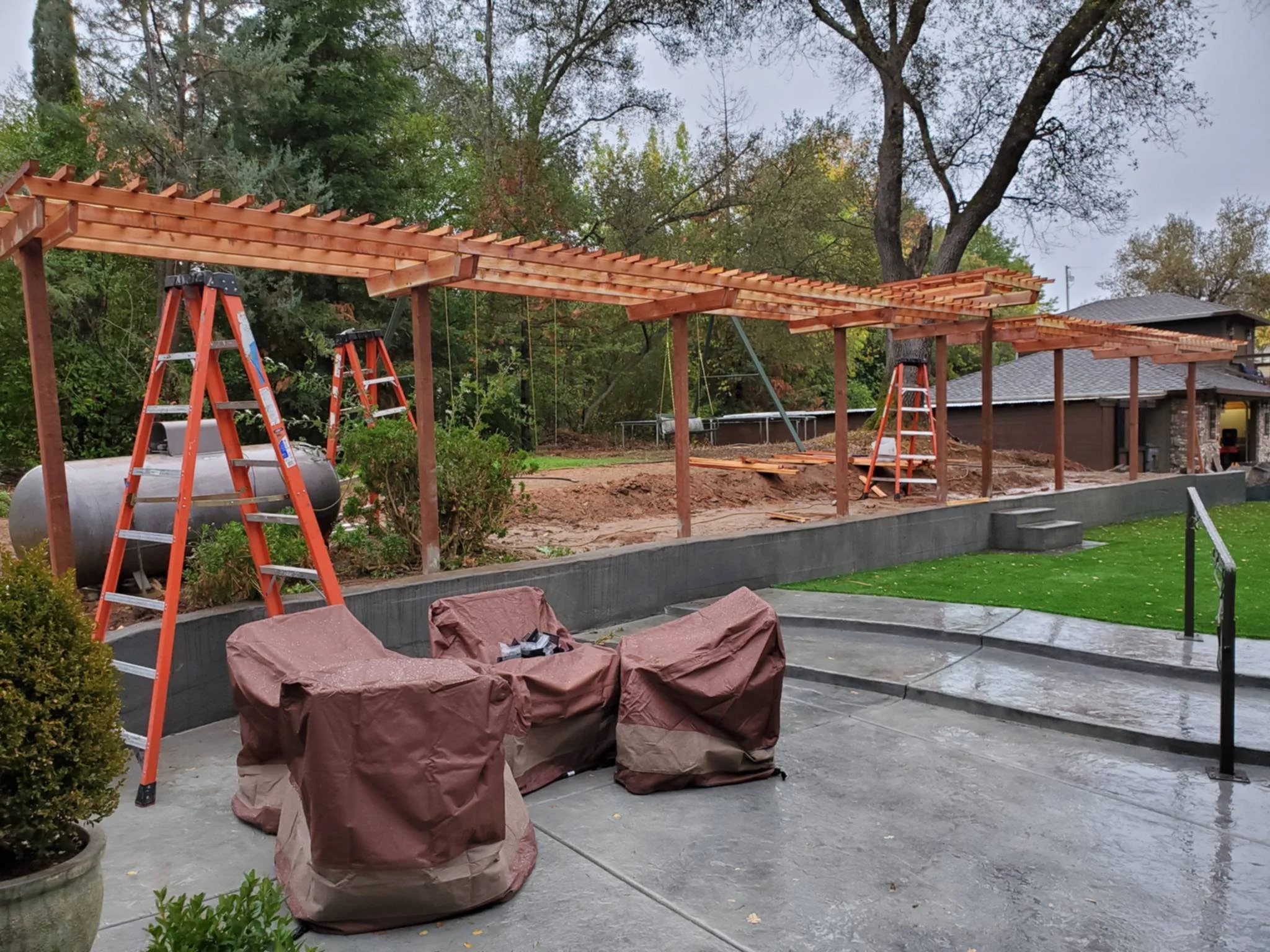 Construction of a wooden pergola in a backyard with ladders and tools, area prepared for additional work, with a grassy lawn and trees in the background.