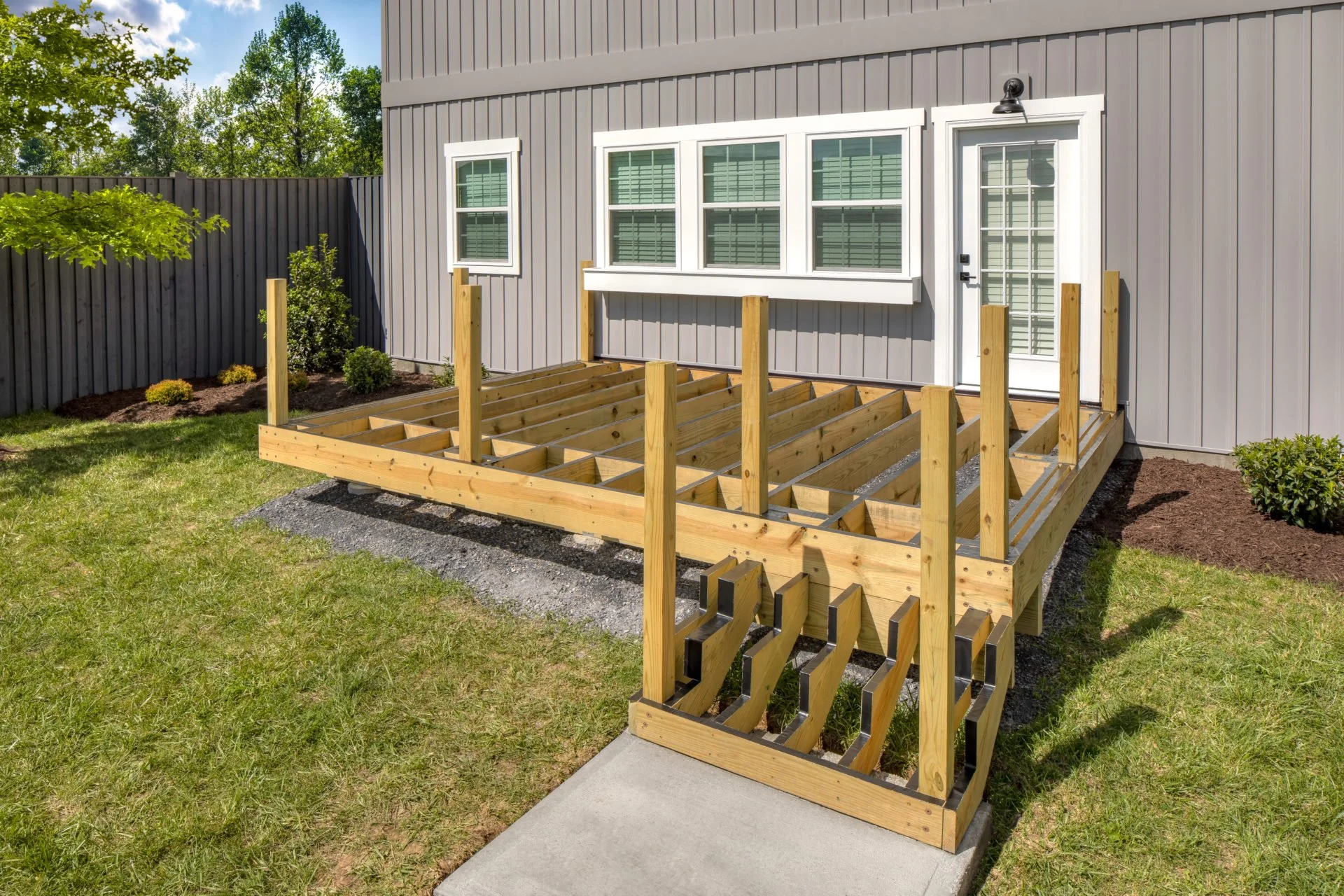 Newly constructed wooden deck with stairs in backyard of gray house with white trim, surrounded by grass, shrubs, and a wooden fence.