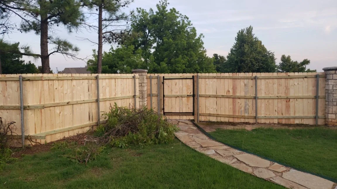 A backyard with a wooden fence, a small gate, a stone pathway, a pile of branches, grass, and trees in the background.