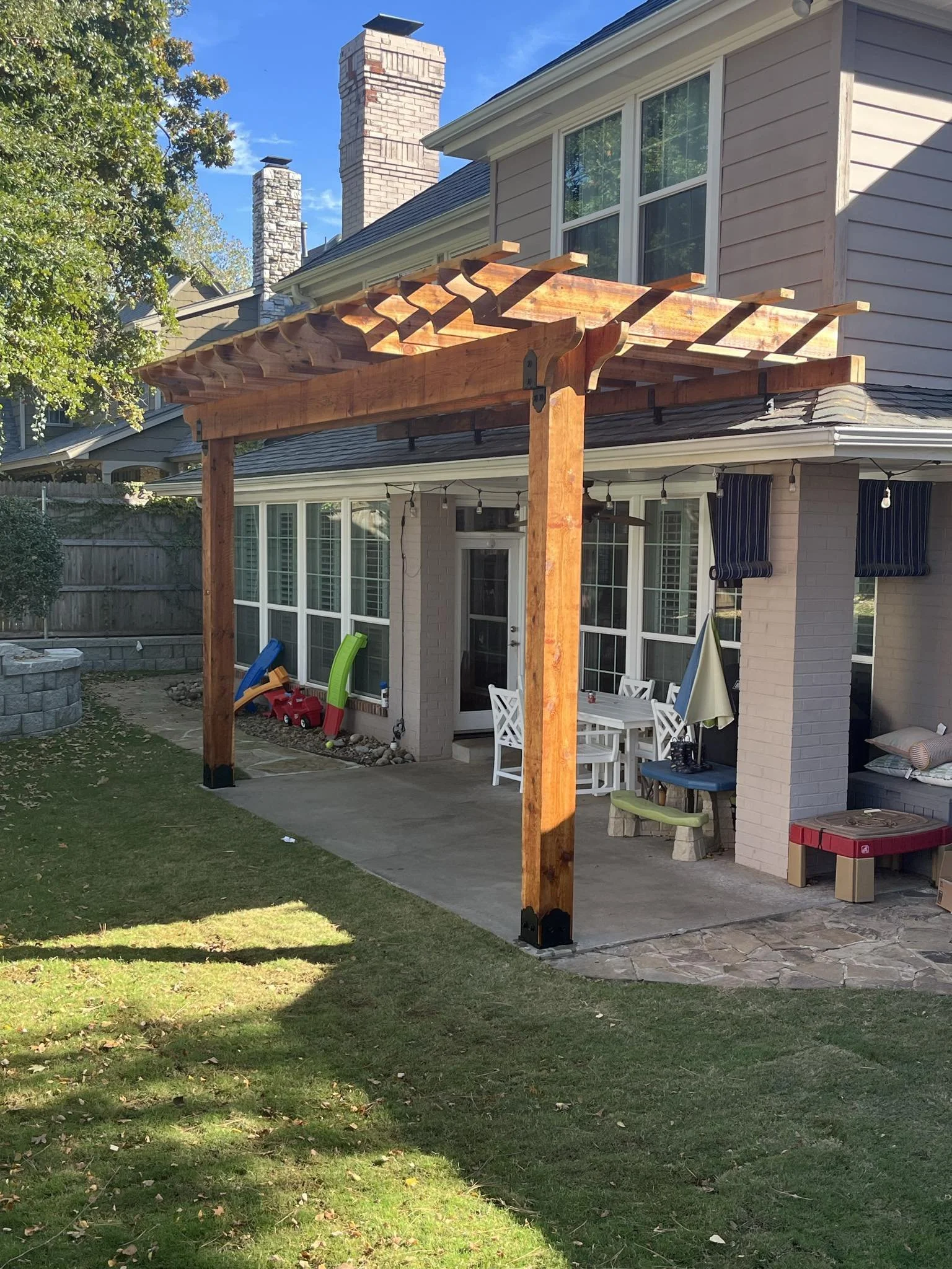 A backyard patio with a newly built wooden pergola, a white outdoor dining table with chairs, a small bench, a red cooler, and children's toys near a glass door of a house with gray siding and multiple chimneys.