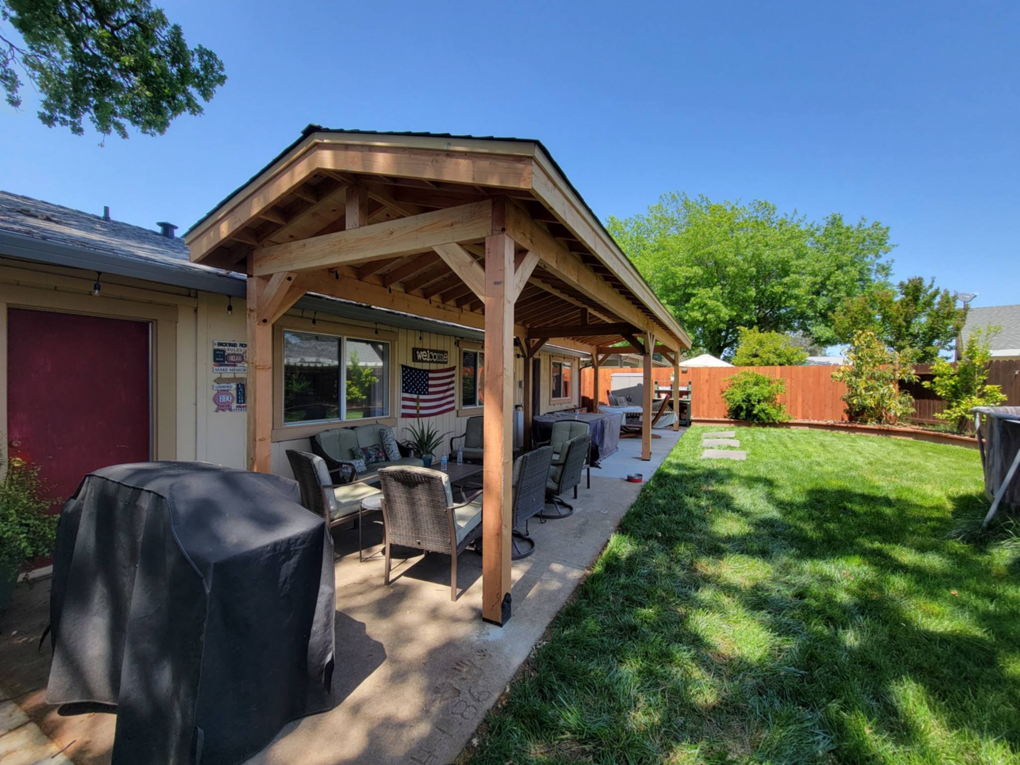 Backyard with a covered patio area, outdoor furniture, a BBQ grill, a well-maintained lawn, trees, and a wooden fence.