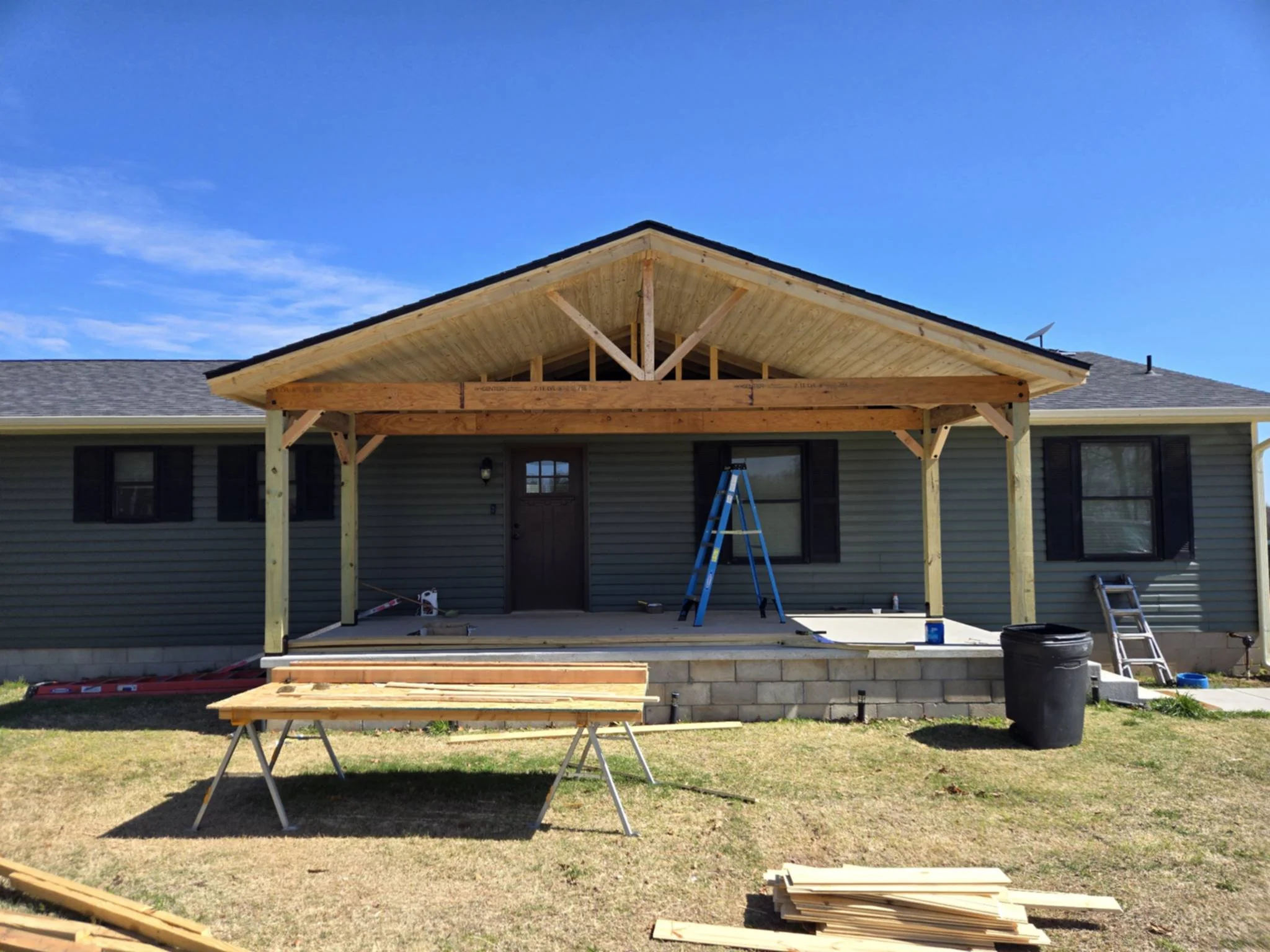 House with a new porch under construction, featuring wooden support beams and a roof extension. Construction tools and materials are visible on the porch and yard.