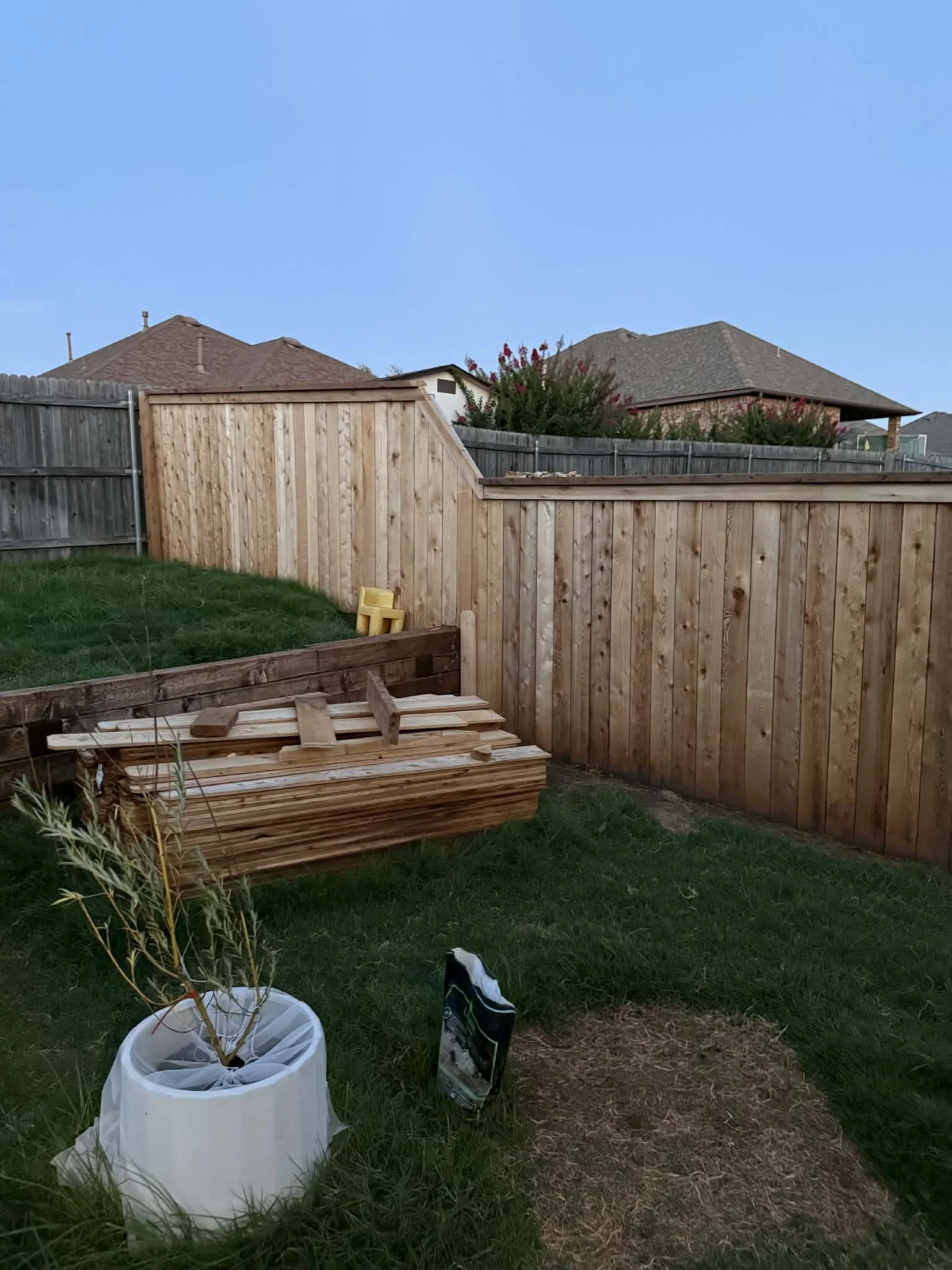 Backyard with newly installed wooden fence, a raised garden bed, a small tree in a white container, and a bag of soil or fertilizer on the ground, with neighboring houses in the background.