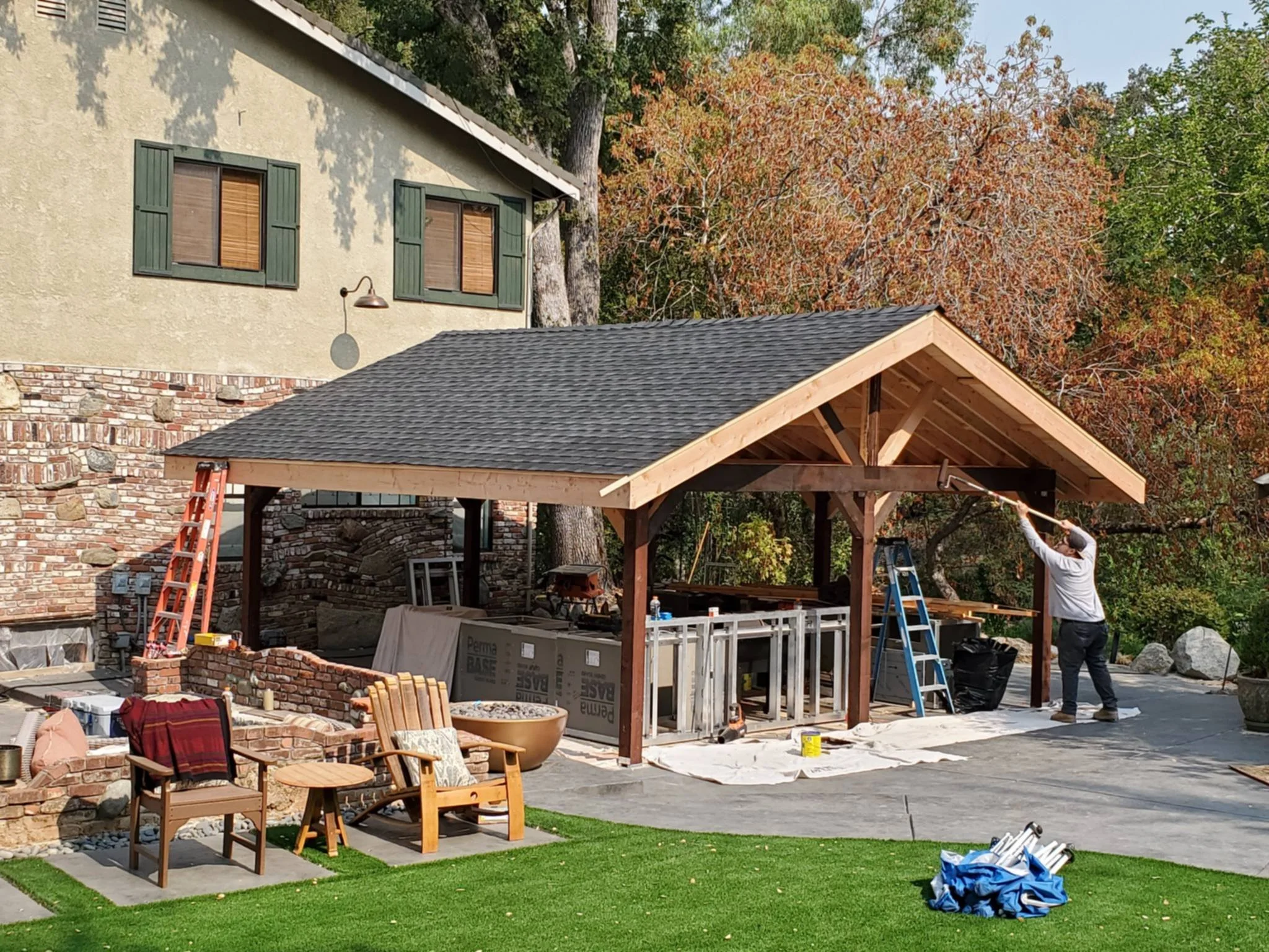 Construction worker painting the underside of a wooden pergola in a backyard with trees and outdoor furniture.