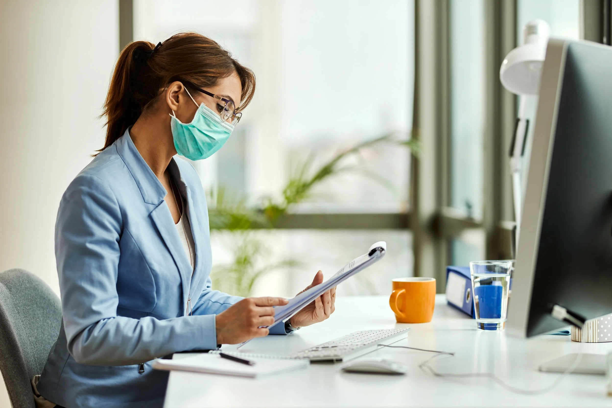 A woman wearing glasses, a blue blazer, and a face mask is sitting at a white desk working with documents, in an office with large windows and plants.