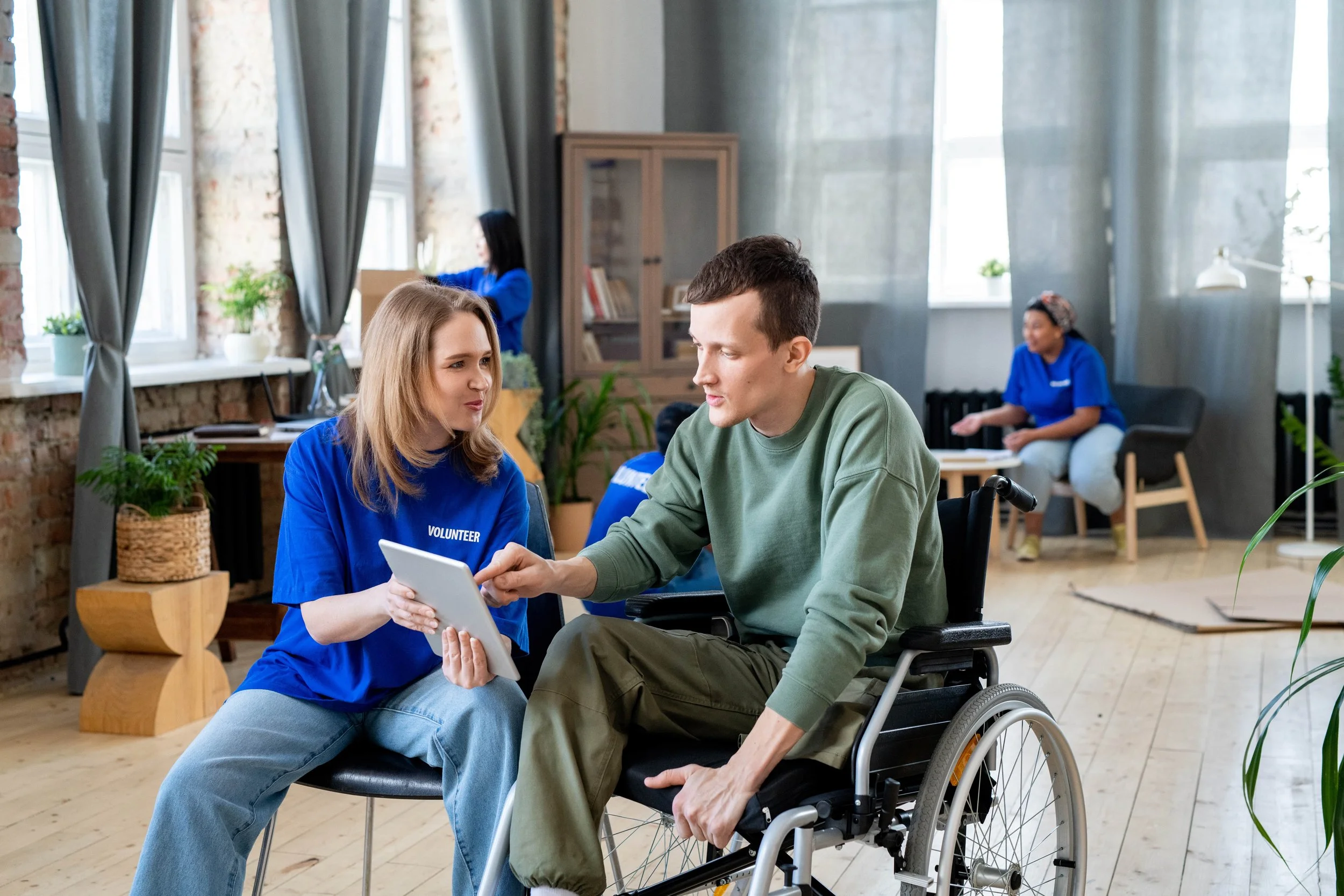 A woman in a blue volunteer shirt helping a man in a green sweatshirt in a wheelchair with a device, in a bright room with coffee tables and large windows.