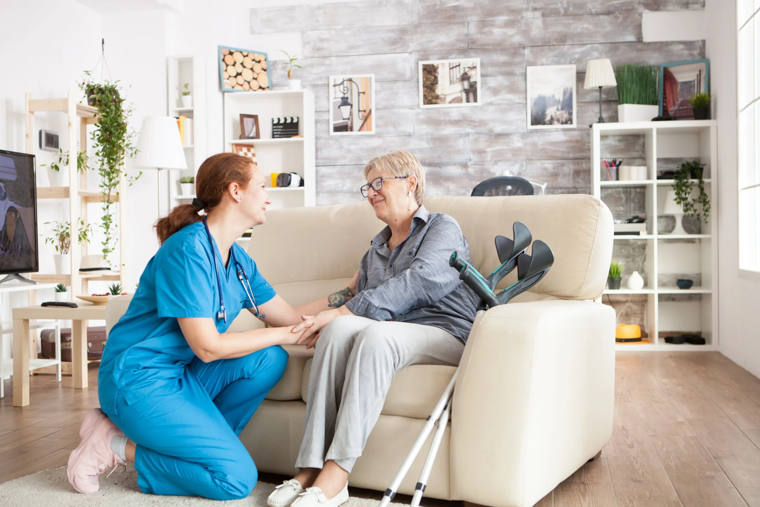 A nurse or caregiver kneeling beside an elderly woman sitting on a beige sofa. The elderly woman, in a wheelchair, is holding the caregiver’s hands and smiling at her. The setting is a cozy living room with modern decor, bookshelves, plants, framed photos, and natural light coming through large windows.