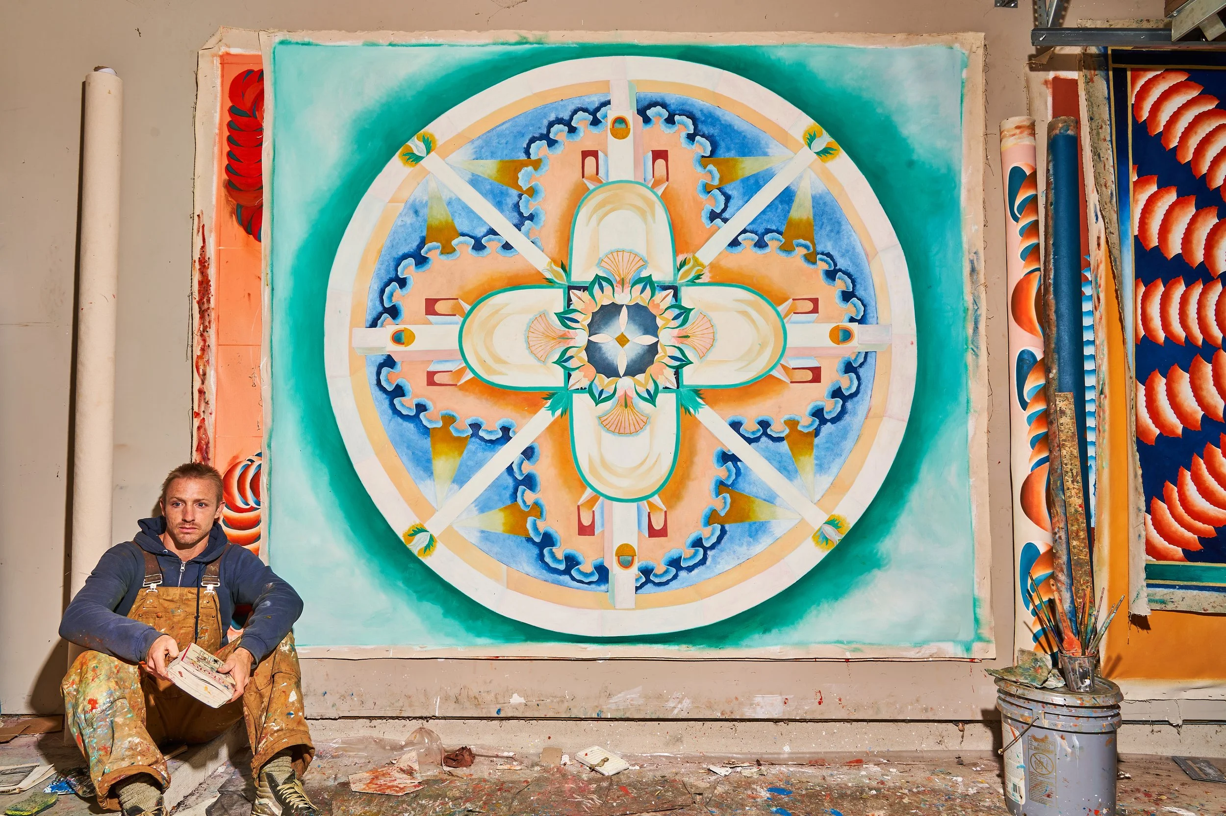 An artist sits on the floor holding a paintbrush in front of a large, colorful, symmetrical abstract painting on a wall. The painting features a central cross shape with floral and geometric patterns in shades of orange, blue, and white, with a green background.