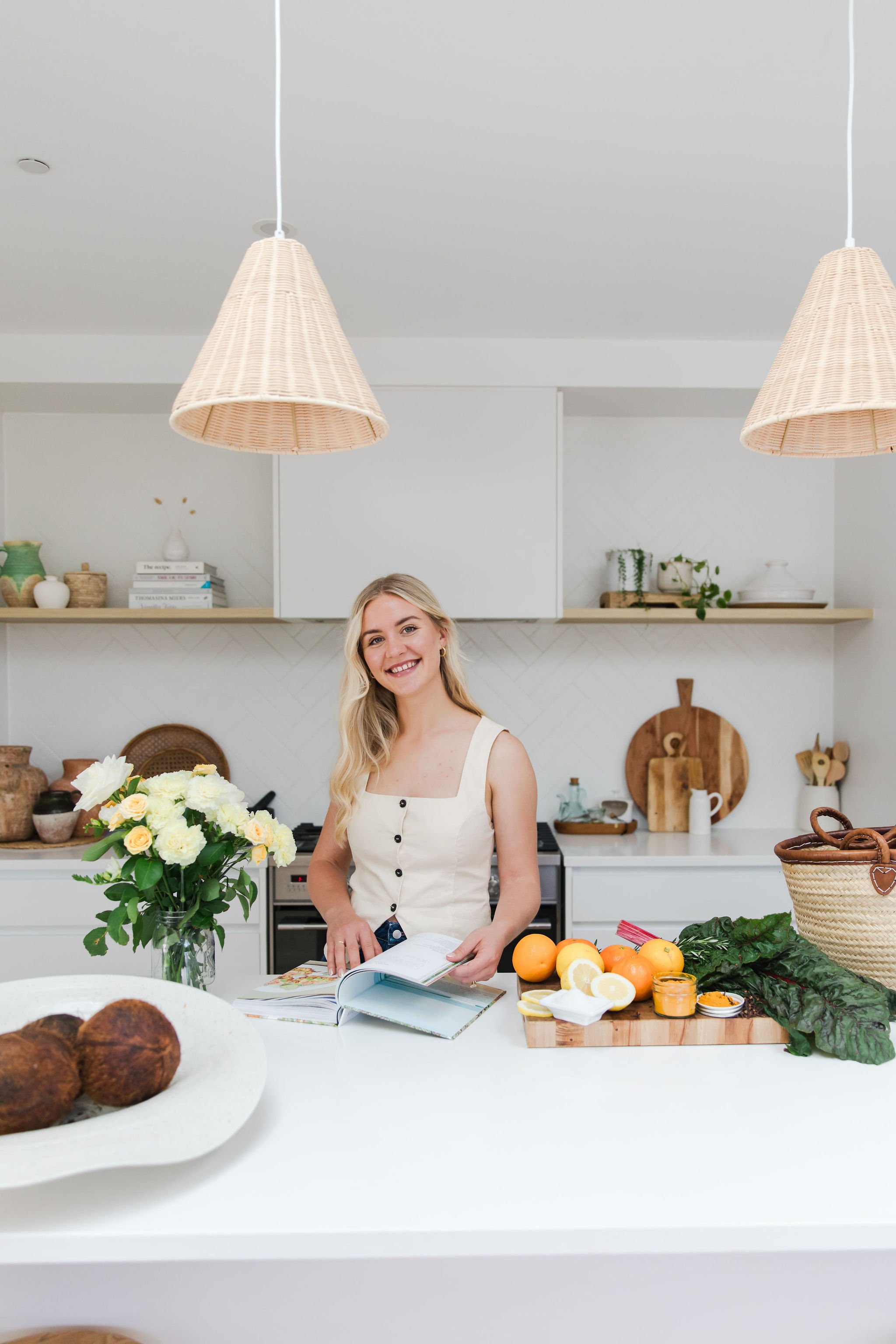 A woman standing in a modern kitchen, smiling, with a book, fresh oranges, lemons, greens, and a vase of white and yellow flowers on a white countertop.