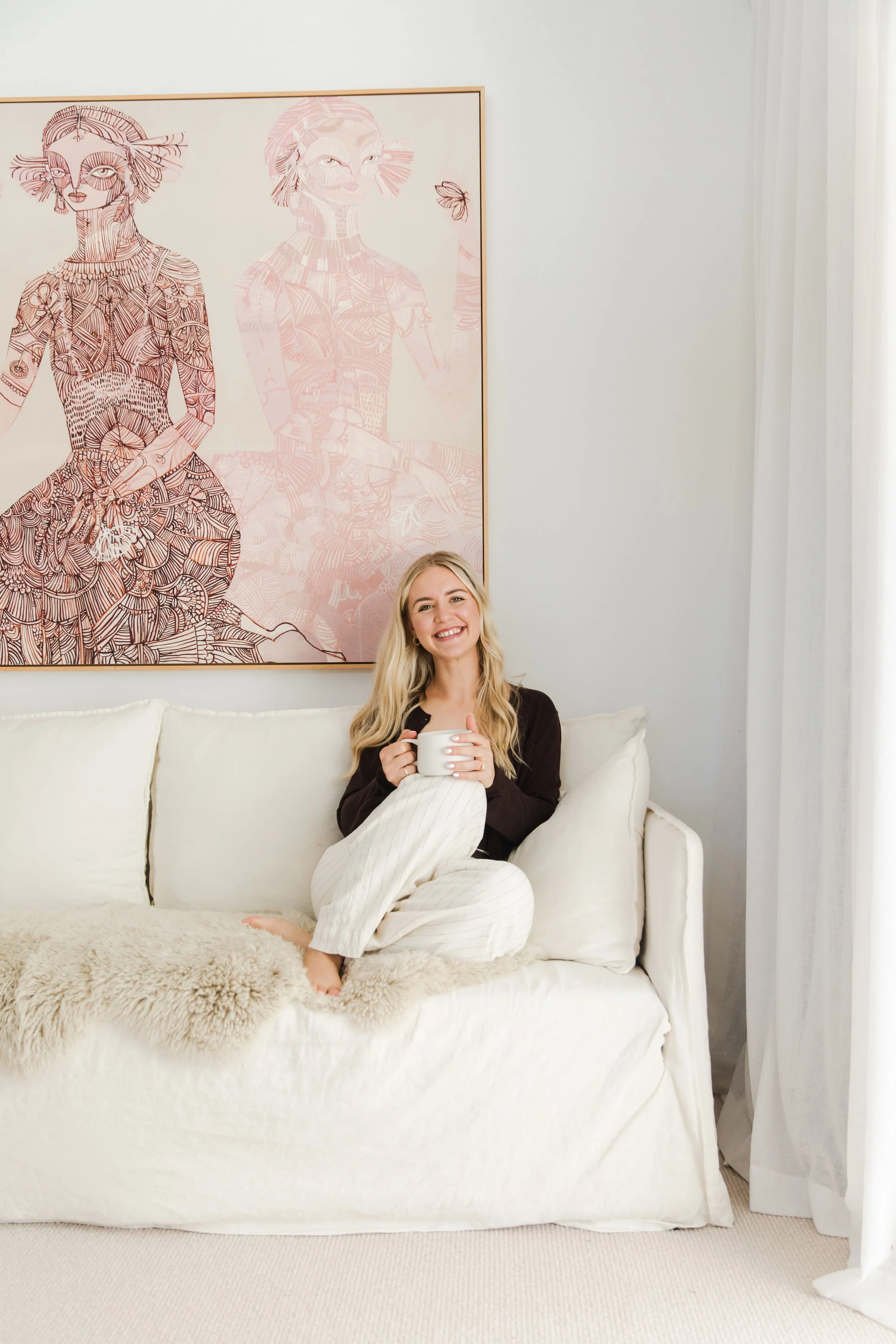 A woman sitting on a white couch holding a mug, smiling, with a large framed artwork behind her featuring an outline of two women with detailed patterned clothing.