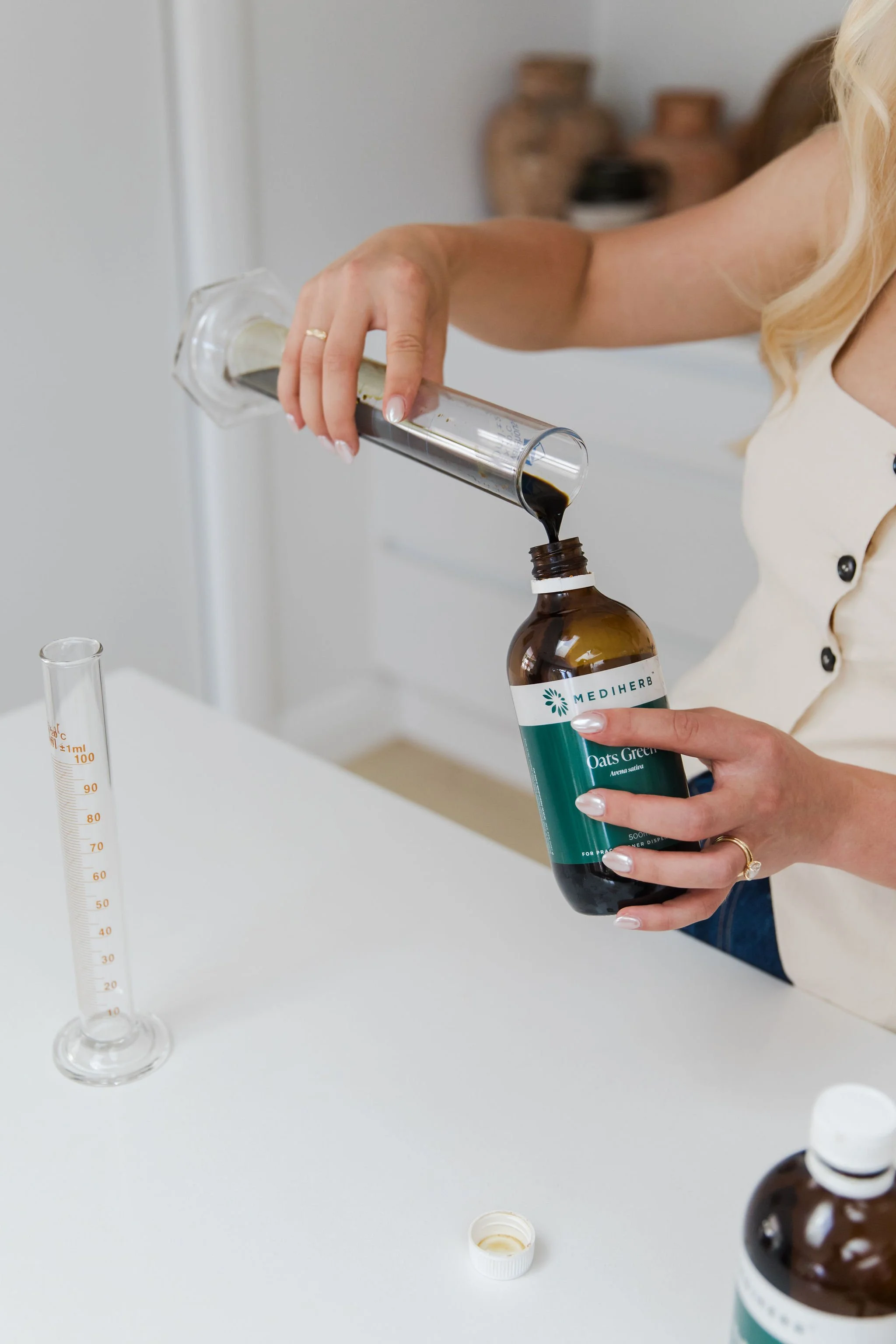 A person pouring a dark liquid from a test tube into a brown bottle labeled 'Mediherb Oats Green' on a white surface, with a graduated measuring cylinder nearby.