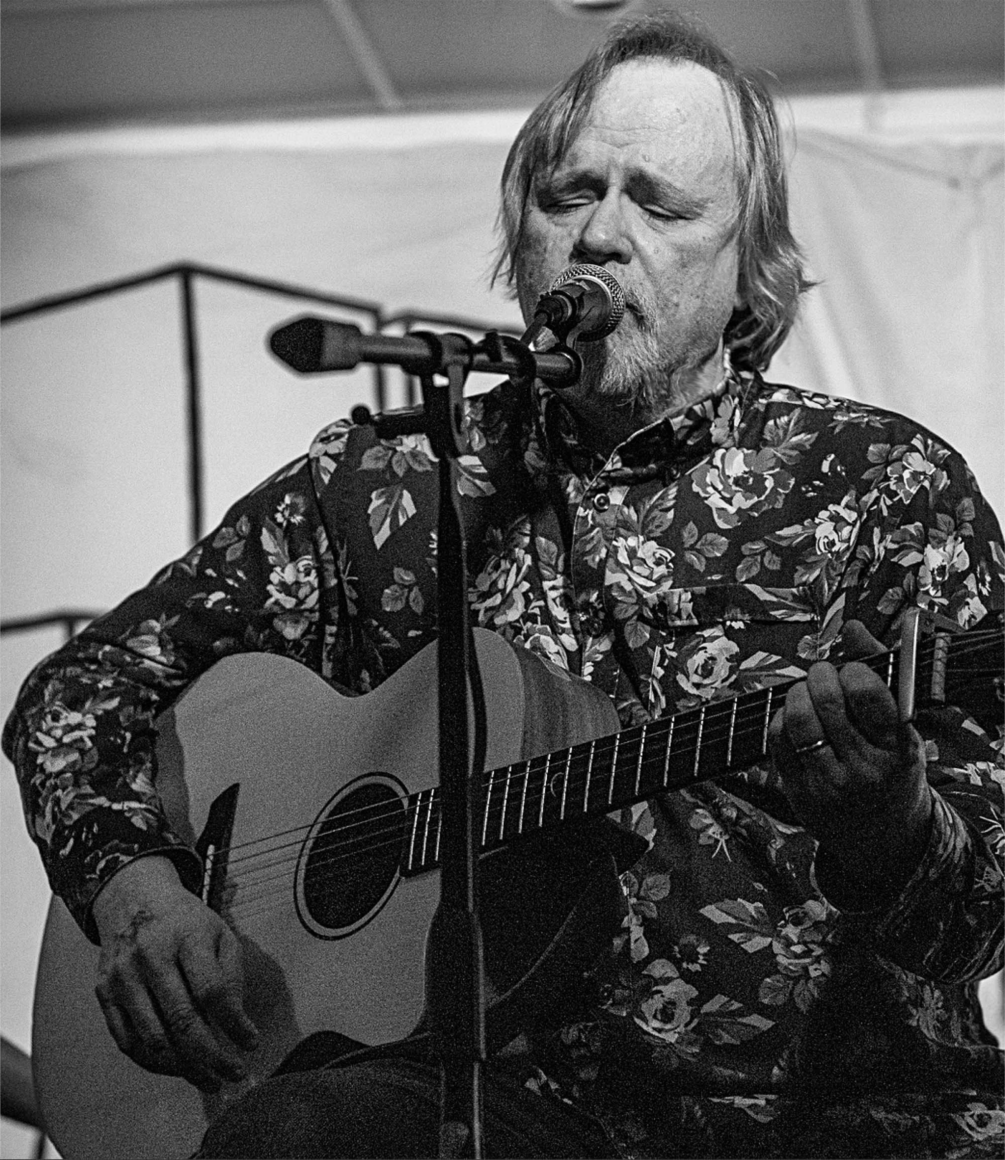 A man with long hair and a beard, wearing a floral shirt, is playing an acoustic guitar and singing into a microphone.