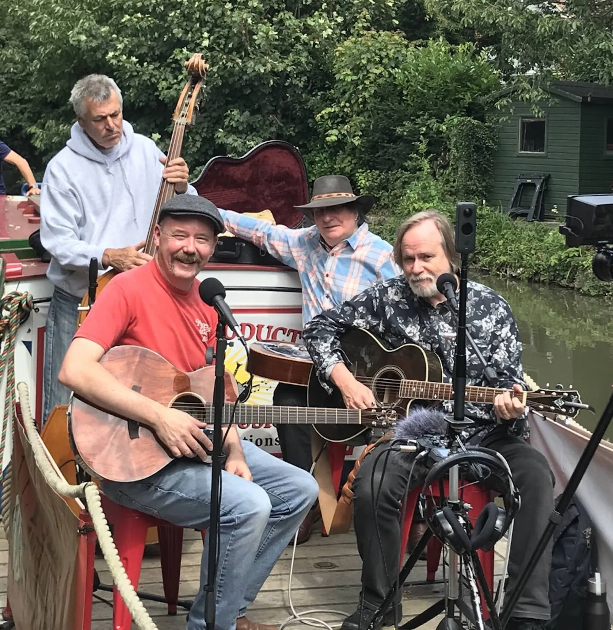 Four men playing musical instruments on a boat, one with a double bass, two with guitars, and one with a microphone, near a body of water with trees and a green house in the background.