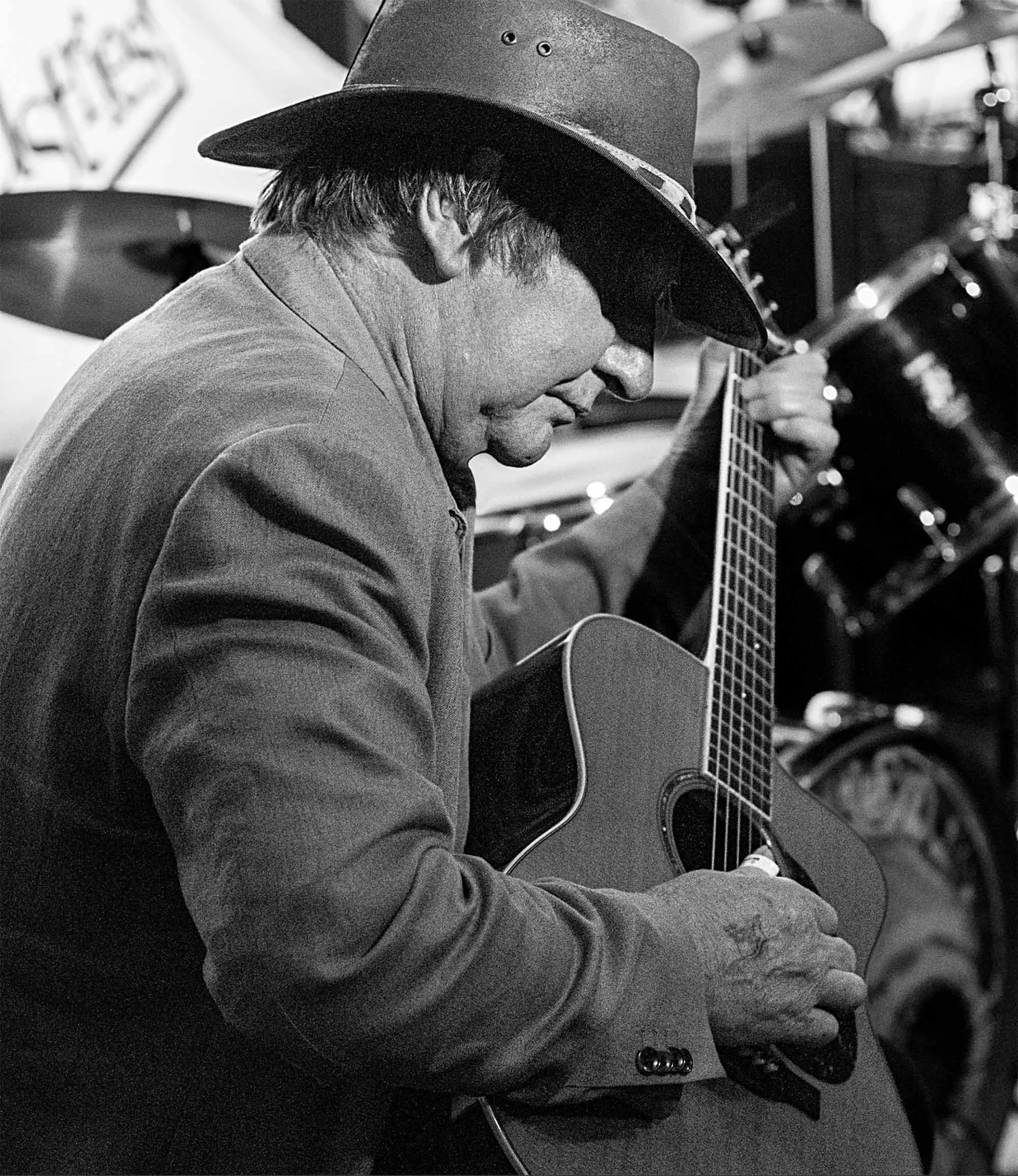 Black and white photo of a man wearing a cowboy hat, playing an acoustic guitar, with a drum set in the background.