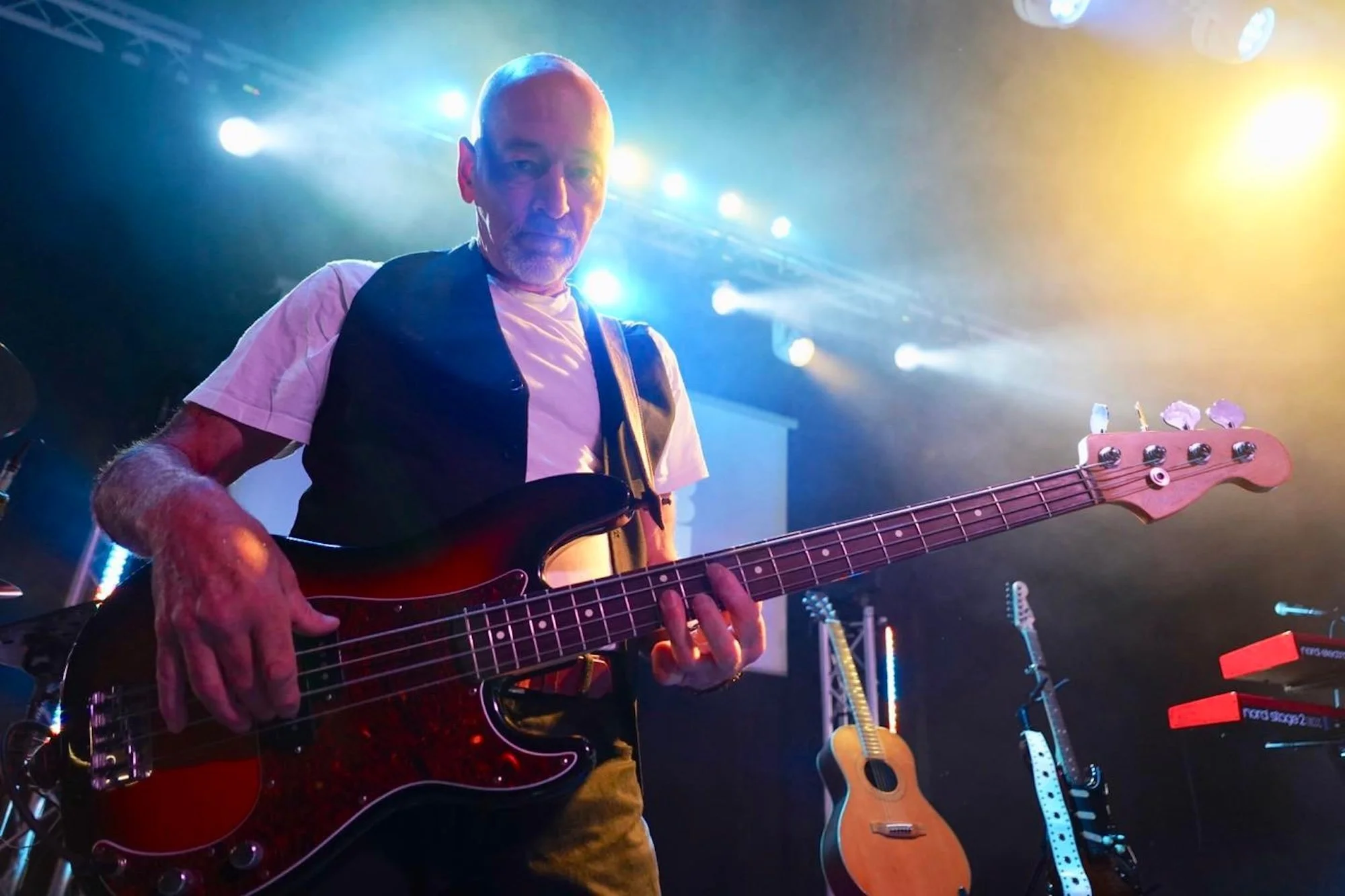 A man playing bass guitar on stage with colorful stage lights, other guitars, and musical equipment visible in the background.