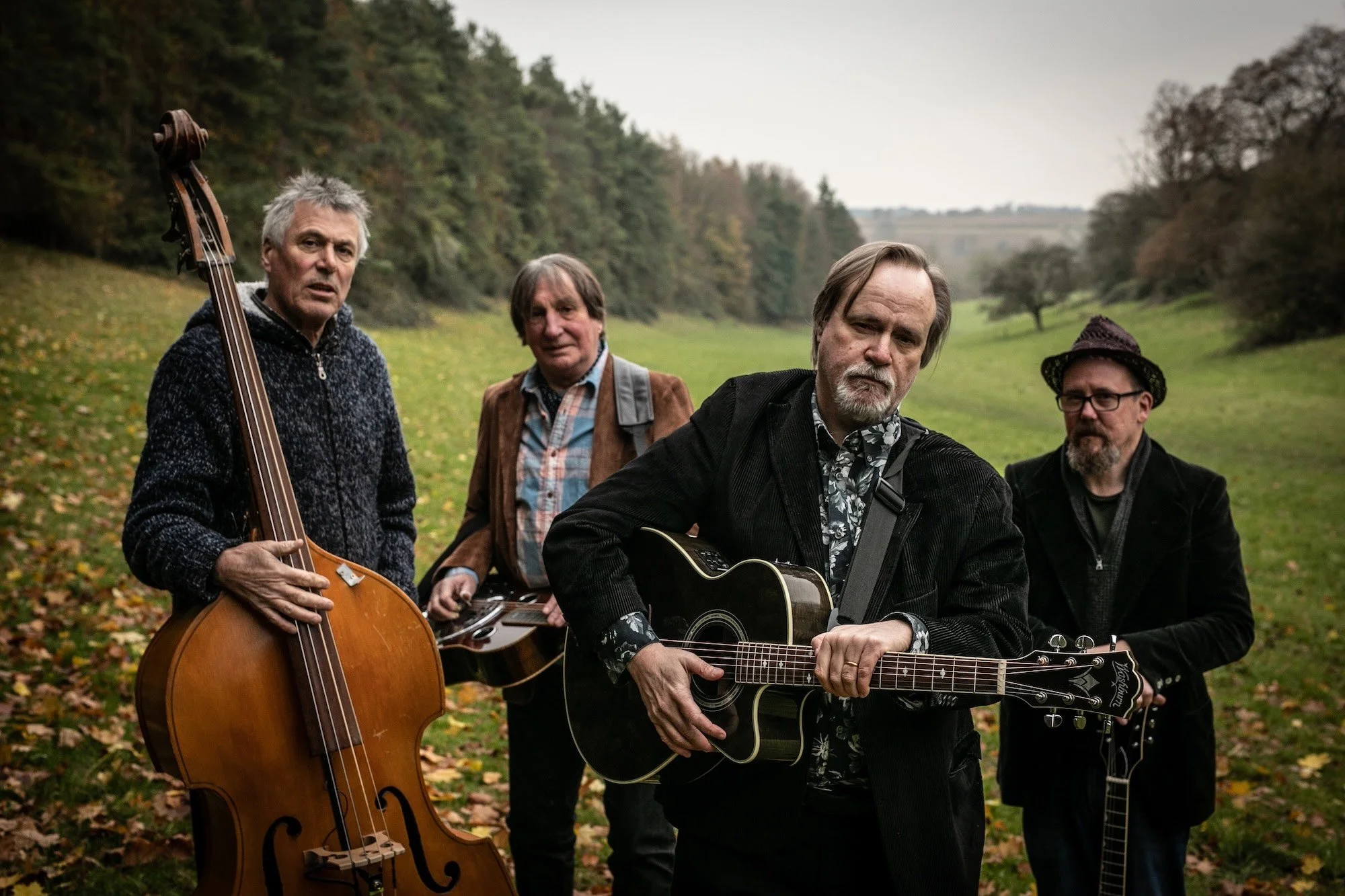 Four men standing outdoors in a grassy field with trees in the background. Two are holding guitars, one with an acoustic guitar and the other with an electric guitar, while another holds a bass guitar. They are dressed in casual, slightly rugged clot