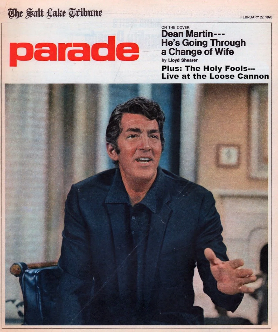 A man with dark, wavy hair, gesturing with his right hand, wearing a dark jacket and over a medium-colored shirt, sitting in a room with family photos on the wall.