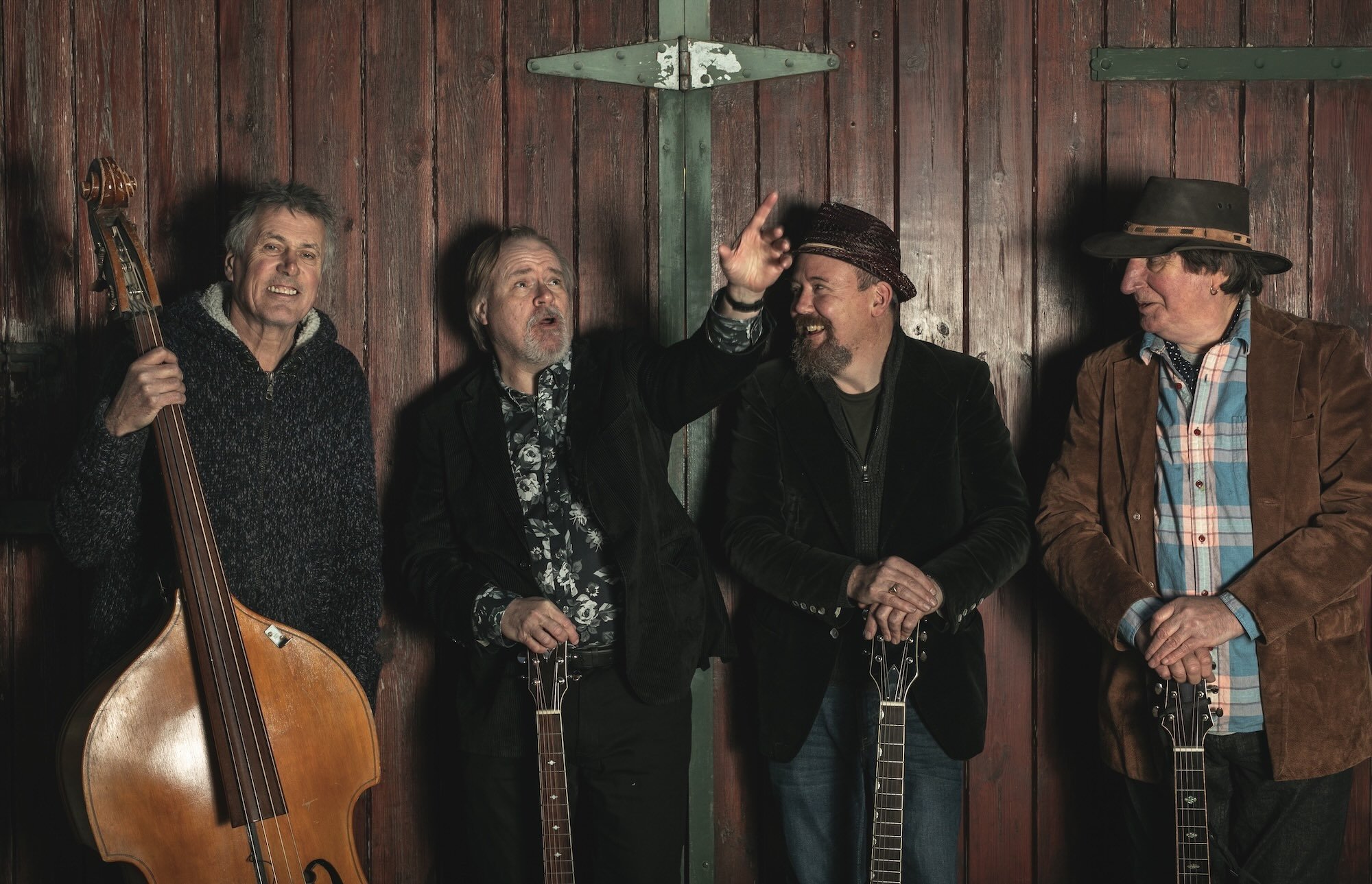 Four men with musical instruments standing in front of a wooden wall, engaging in conversation and smiling.