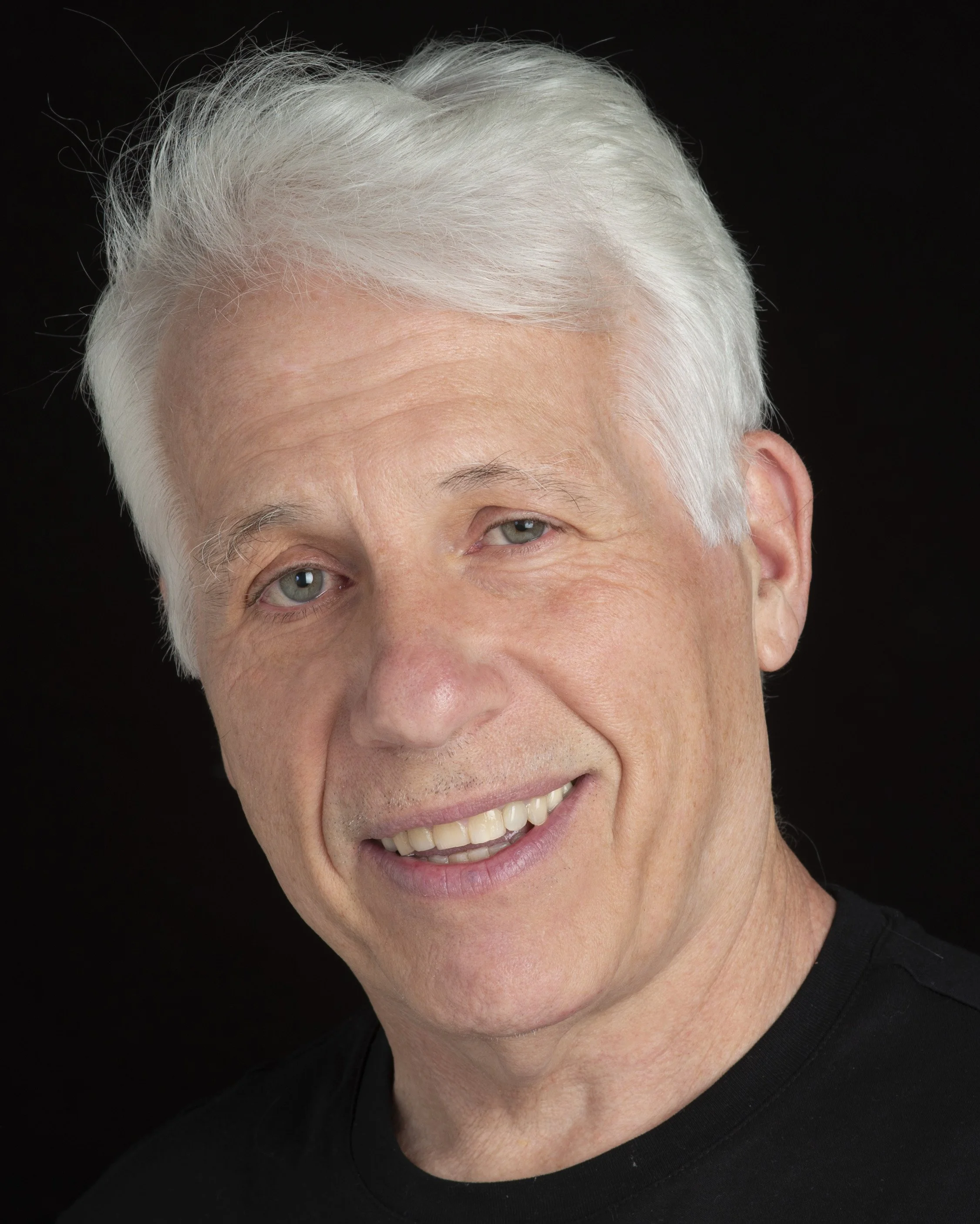 A smiling older man with white hair, blue eyes, and light skin, wearing a black shirt against a black background.