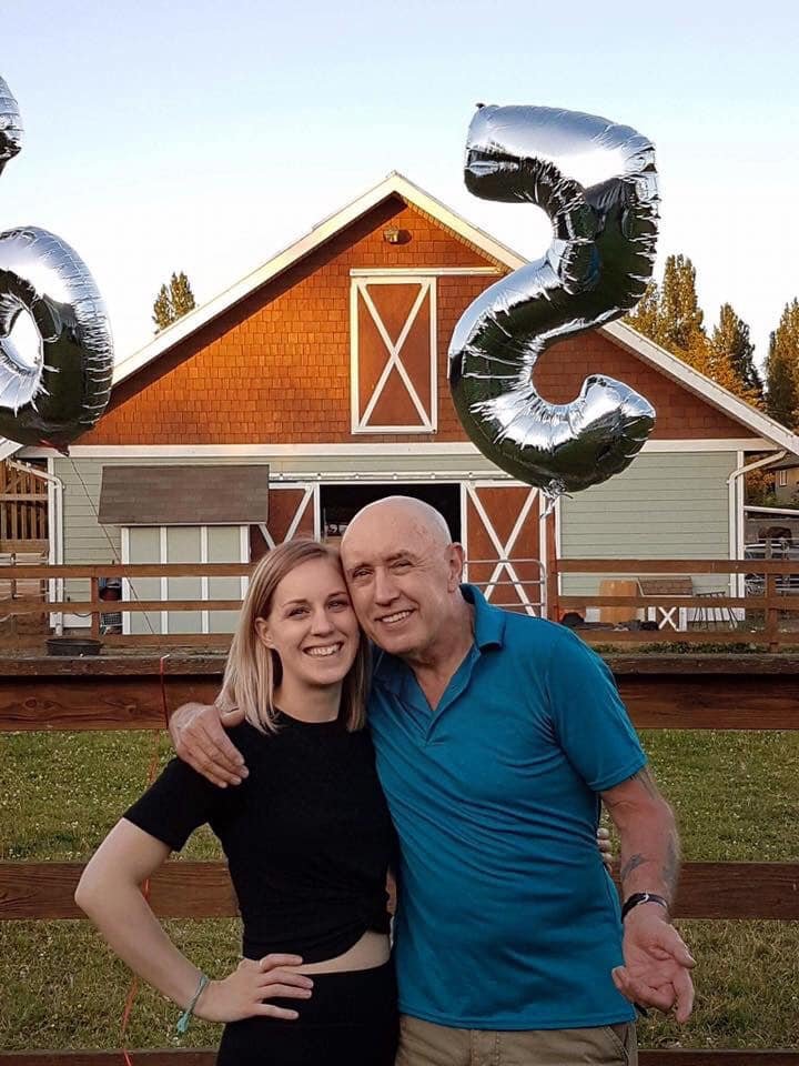 A young woman and an older man smiling and hugging in front of a wooden fence with a barn in the background. There are black helium balloons shaped as the number 2 and 3, indicating a celebration of a 23rd milestone.