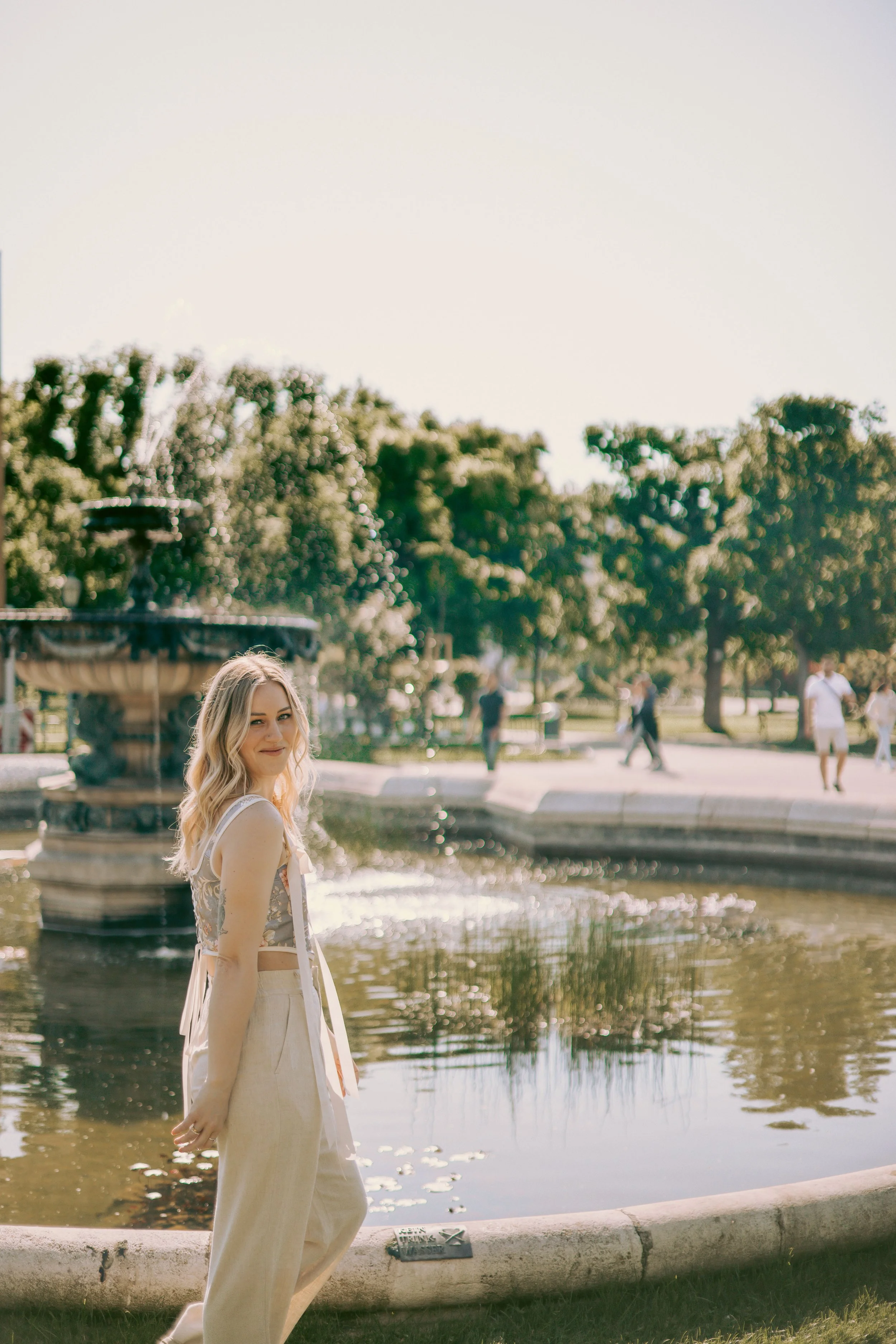 A woman with blonde hair in a garden near a fountain with water, trees, and people walking in the background.