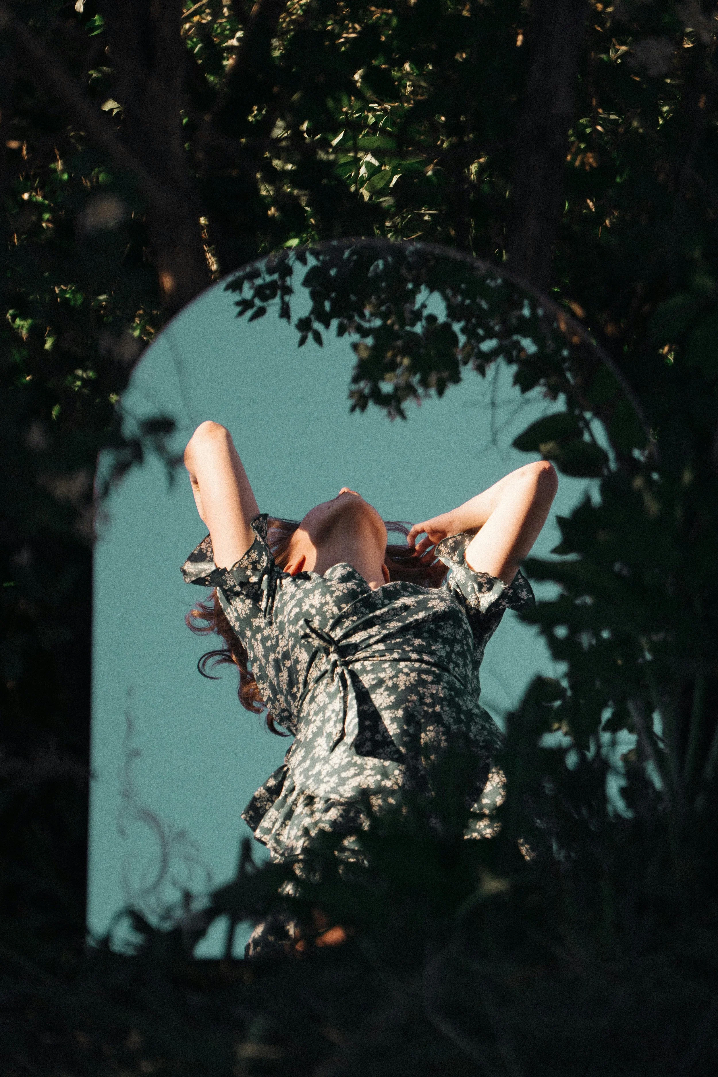 A woman with curly brown hair, her face partly visible, is posing outdoors in front of trees with green leaves. She is wearing a black floral dress with ruffled sleeves and has her arms raised behind her head.