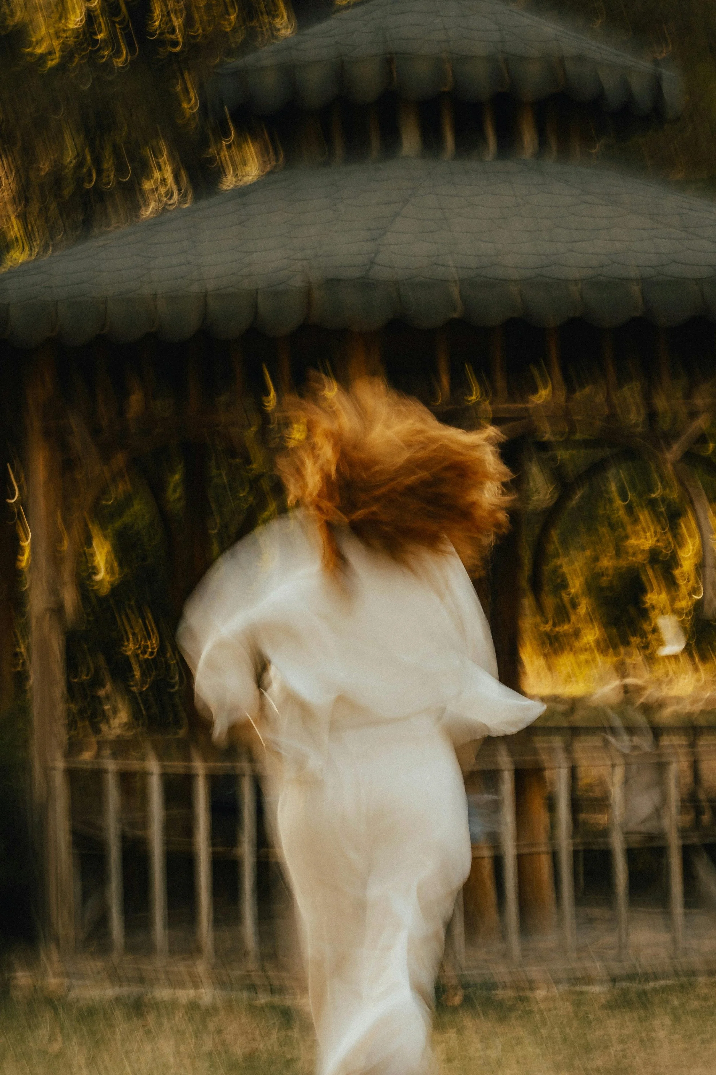 Blurred image showing a person in white clothing with reddish-brown hair walking past a metal fence at night, with warm lights in the background.