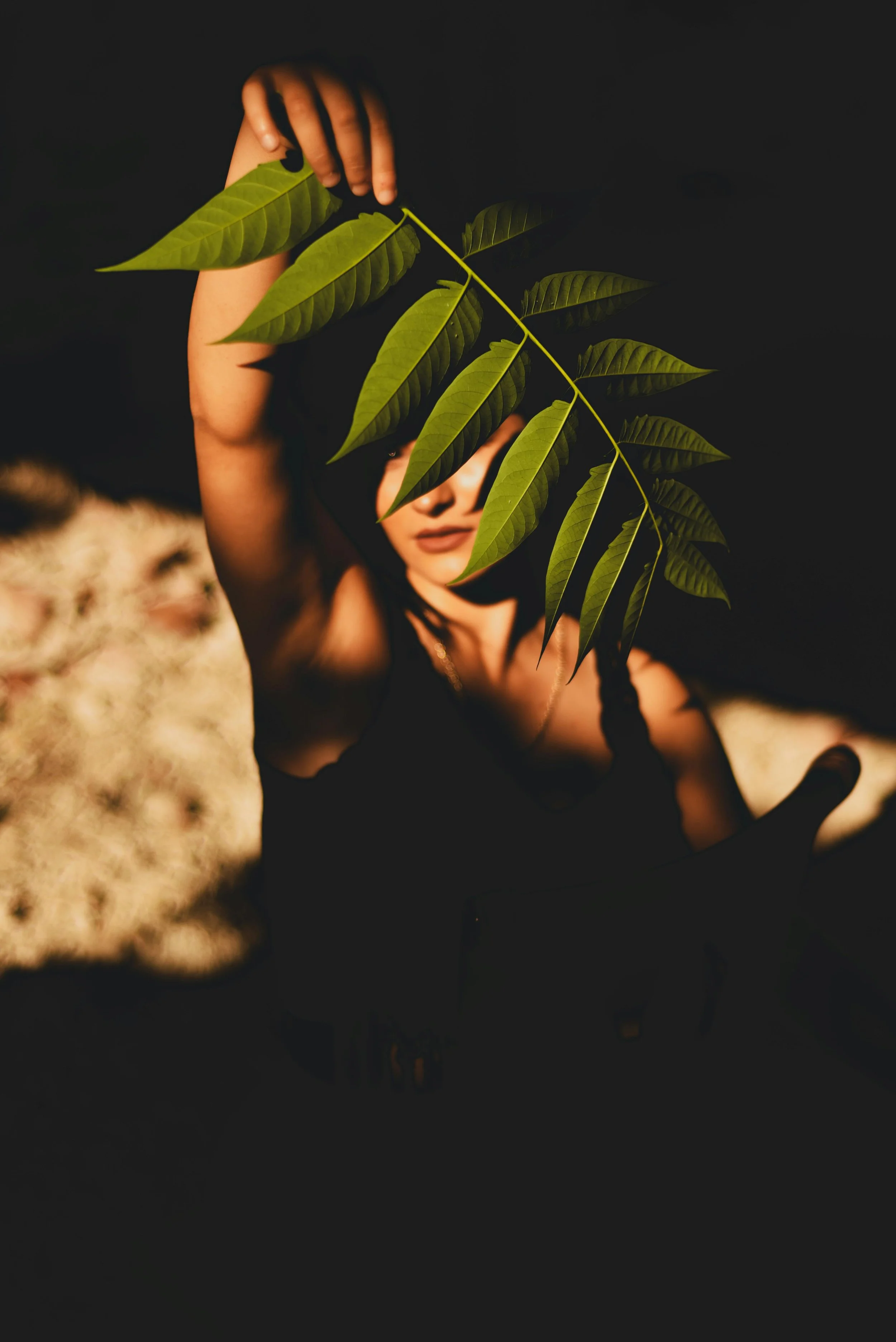 A woman holding a large green leafy branch in front of her face, with dark background and golden brown hair.