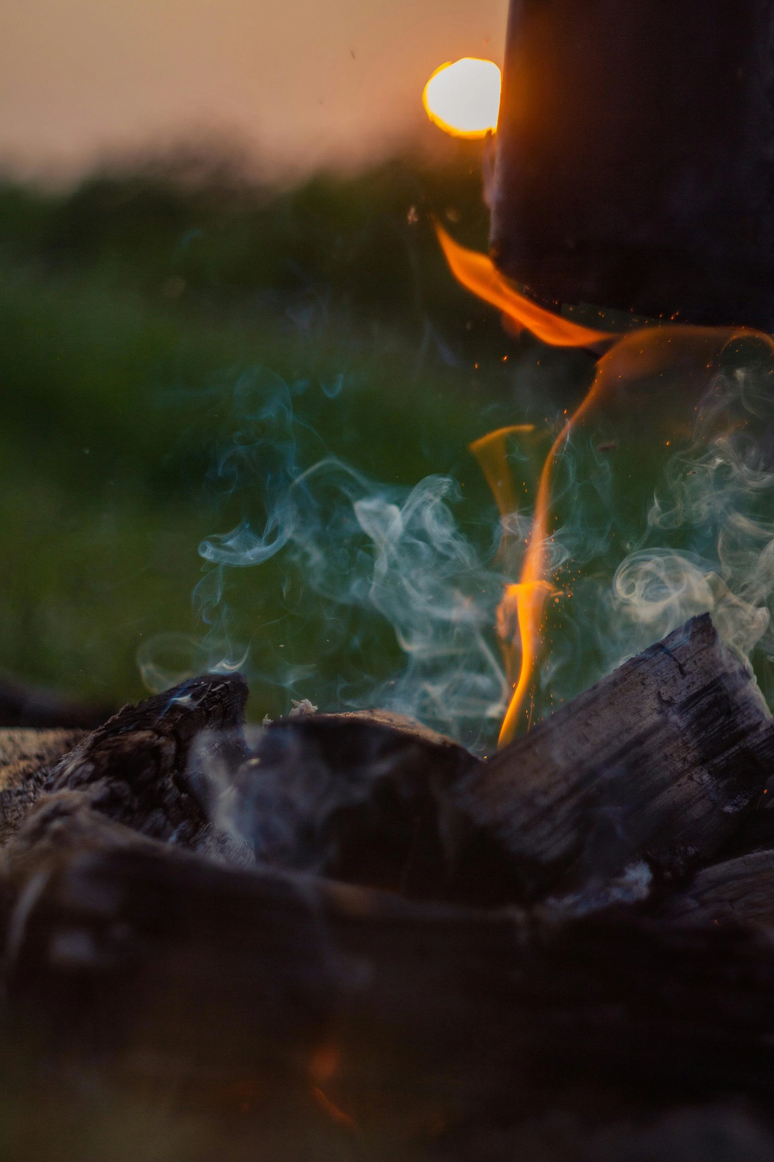 Close-up of a campfire with glowing orange flames and smoke. Logs are visible at the base of the fire.