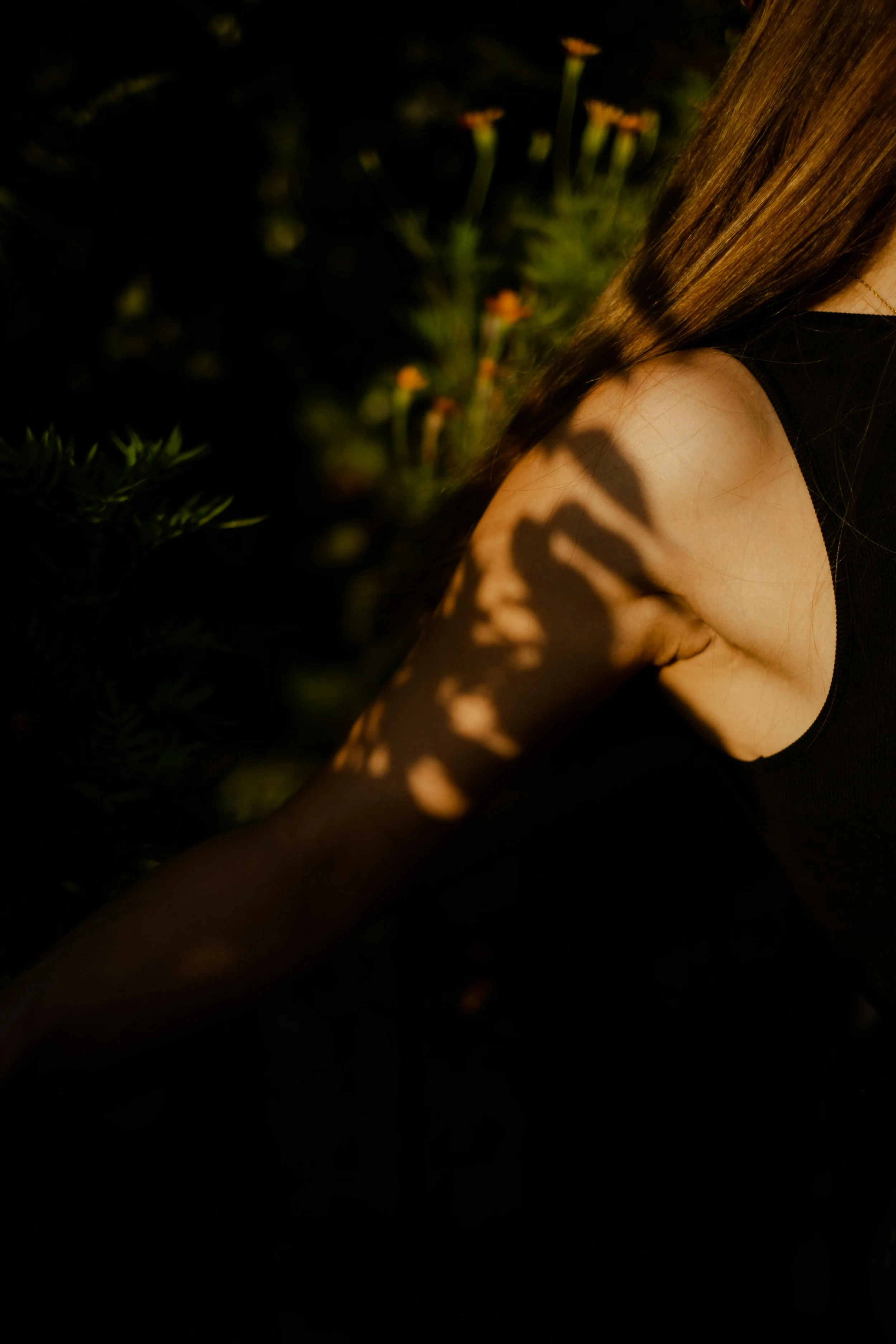 Close-up of a woman's shoulder with long hair, partially covered in shadows from leaves, set against a dark background with faintly visible flowers or plants.