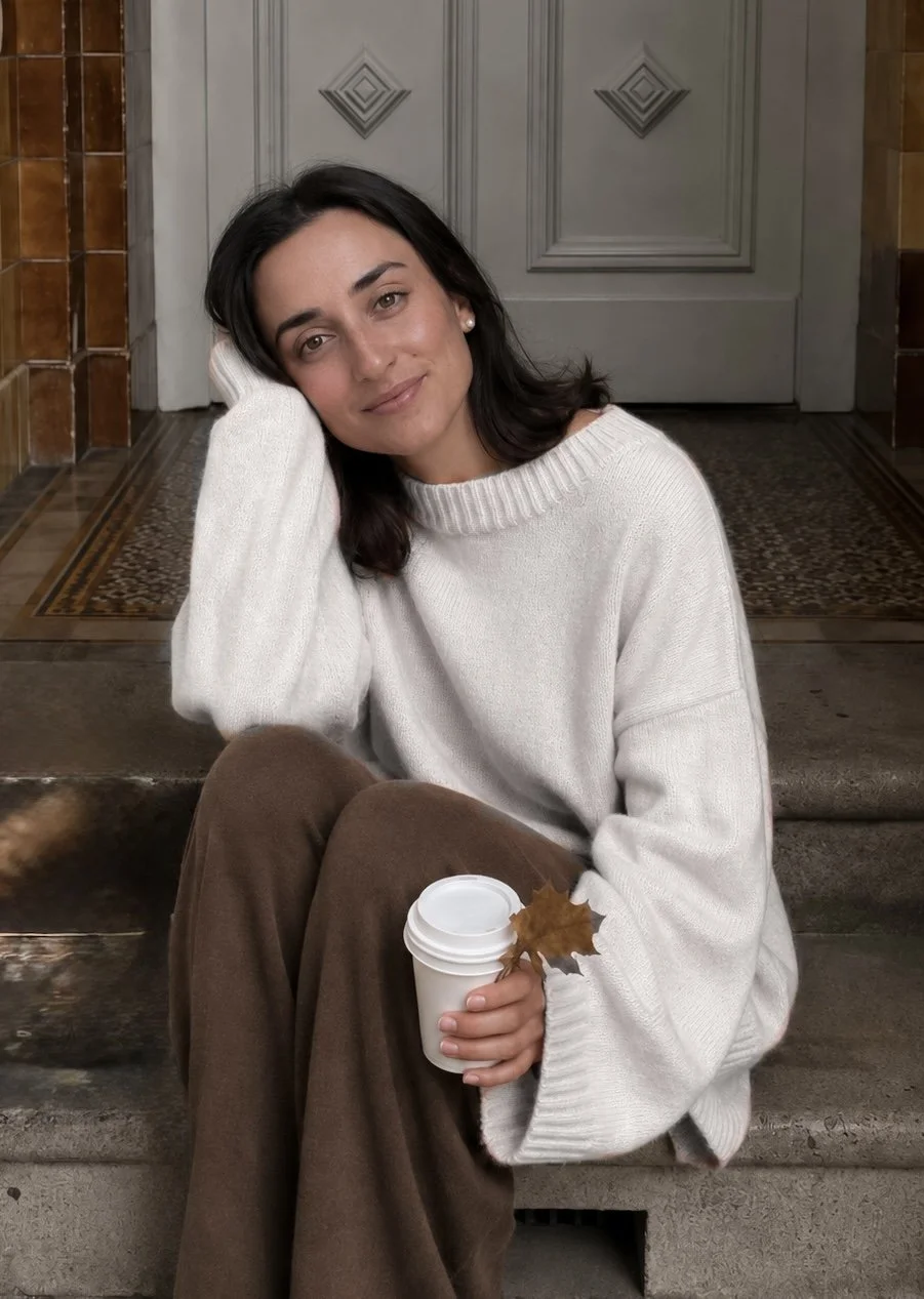 Una donna seduta sulle scale in un ambiente interno, che sorseggia una bevanda in una tazza di carta, con una foglia d'acero. Indossa un maglione bianco e pantaloni marroni, con uno sfondo di porta e pareti decorate.