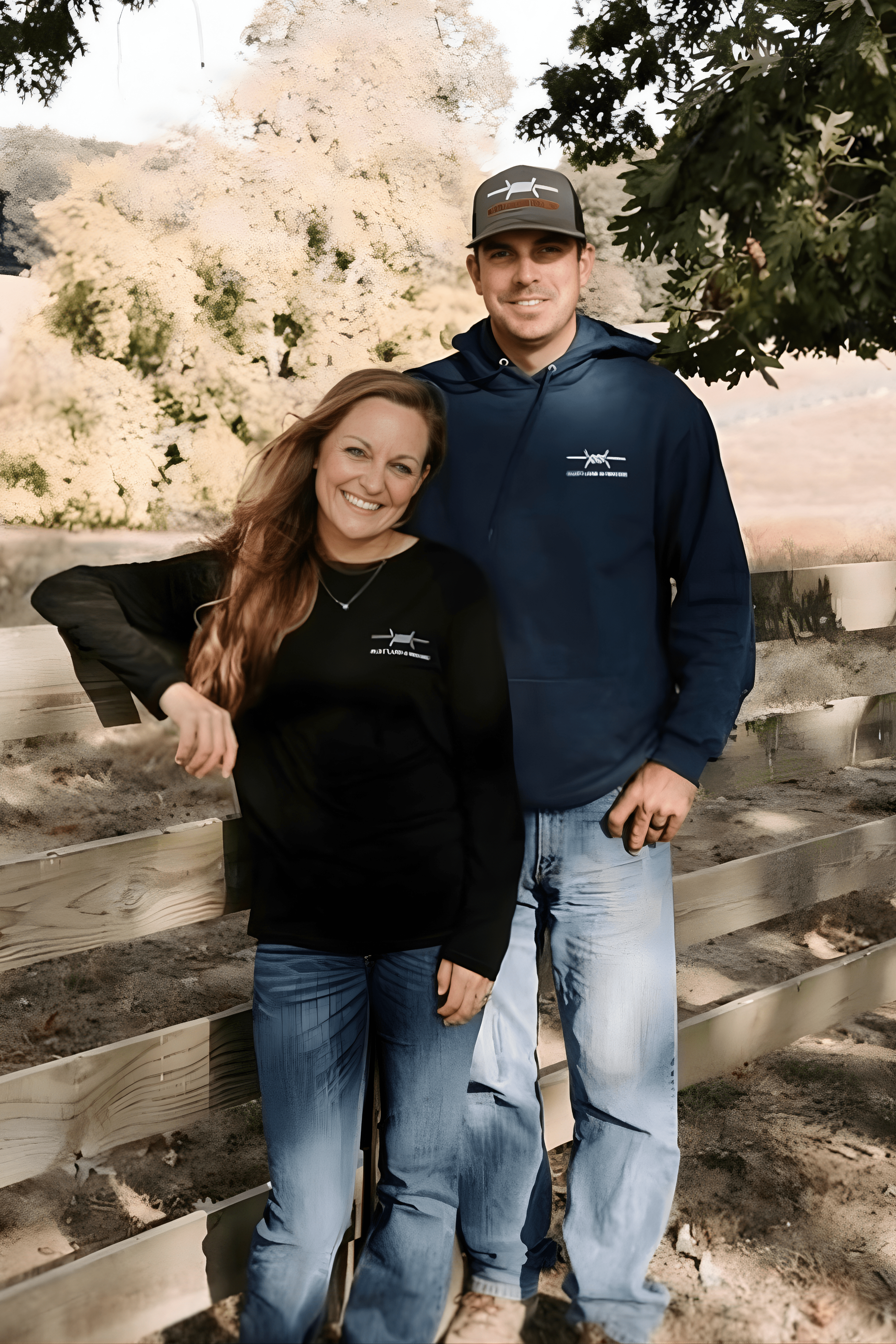 A smiling woman and man stand outside near a wooden fence with trees and white flowers in the background, both wearing dark hoodies with airplane logos.