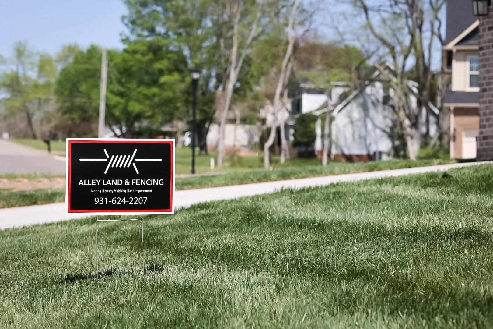 Sign for Alley Land & Fencing placed in a front yard, with a suburban street and trees in the background.